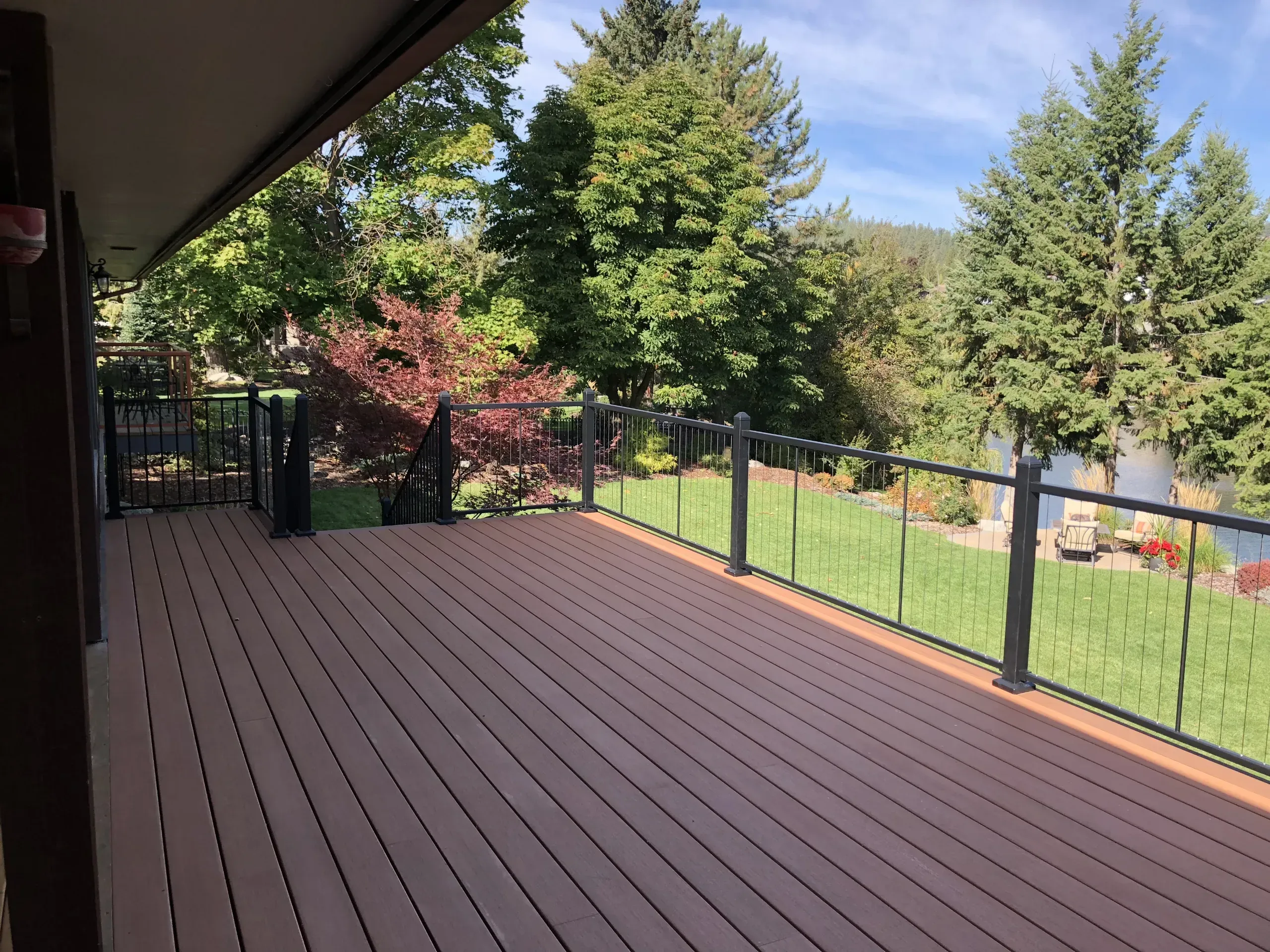 A wide composite deck in full sun with black cable railing, looking out over a mown lawn, mature trees and a river beyond.