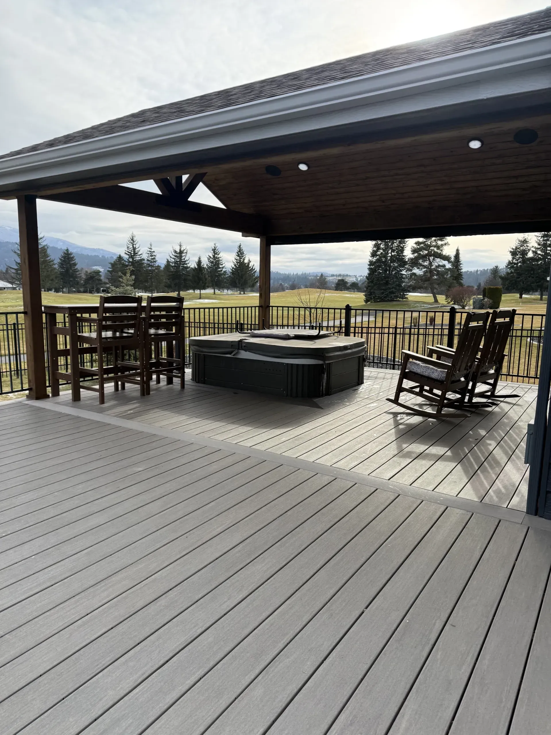 A covered composite deck with a hot tub, bar seating and rocking chairs, looking out over open fairway and mountains.
