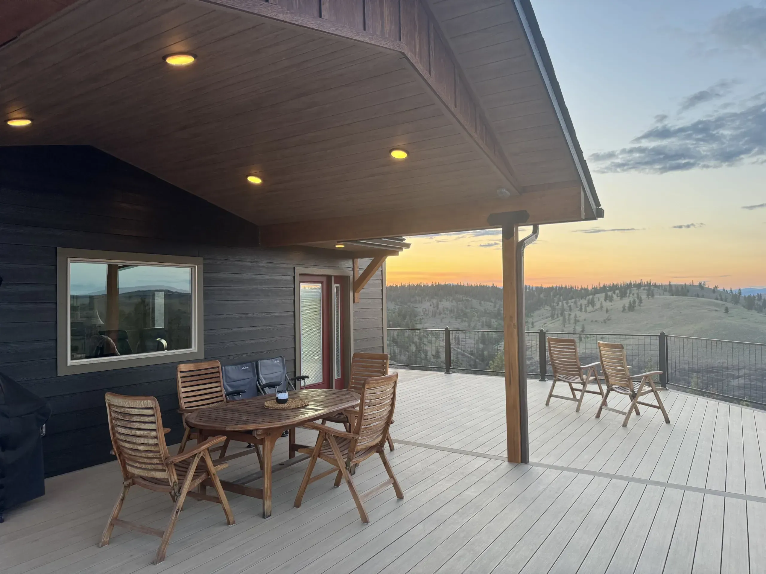 A covered composite deck at sunset, its cedar ceiling lit by recessed downlights, with cable railing looking out over rolling hills.