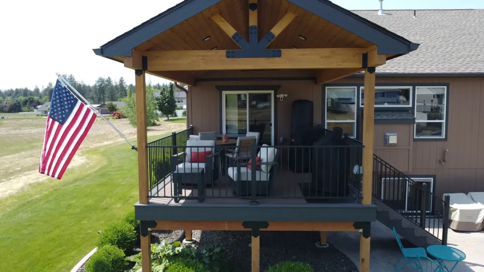 A raised deck under a gabled cedar cover with exposed beams, furnished with outdoor seating and a dining table.
