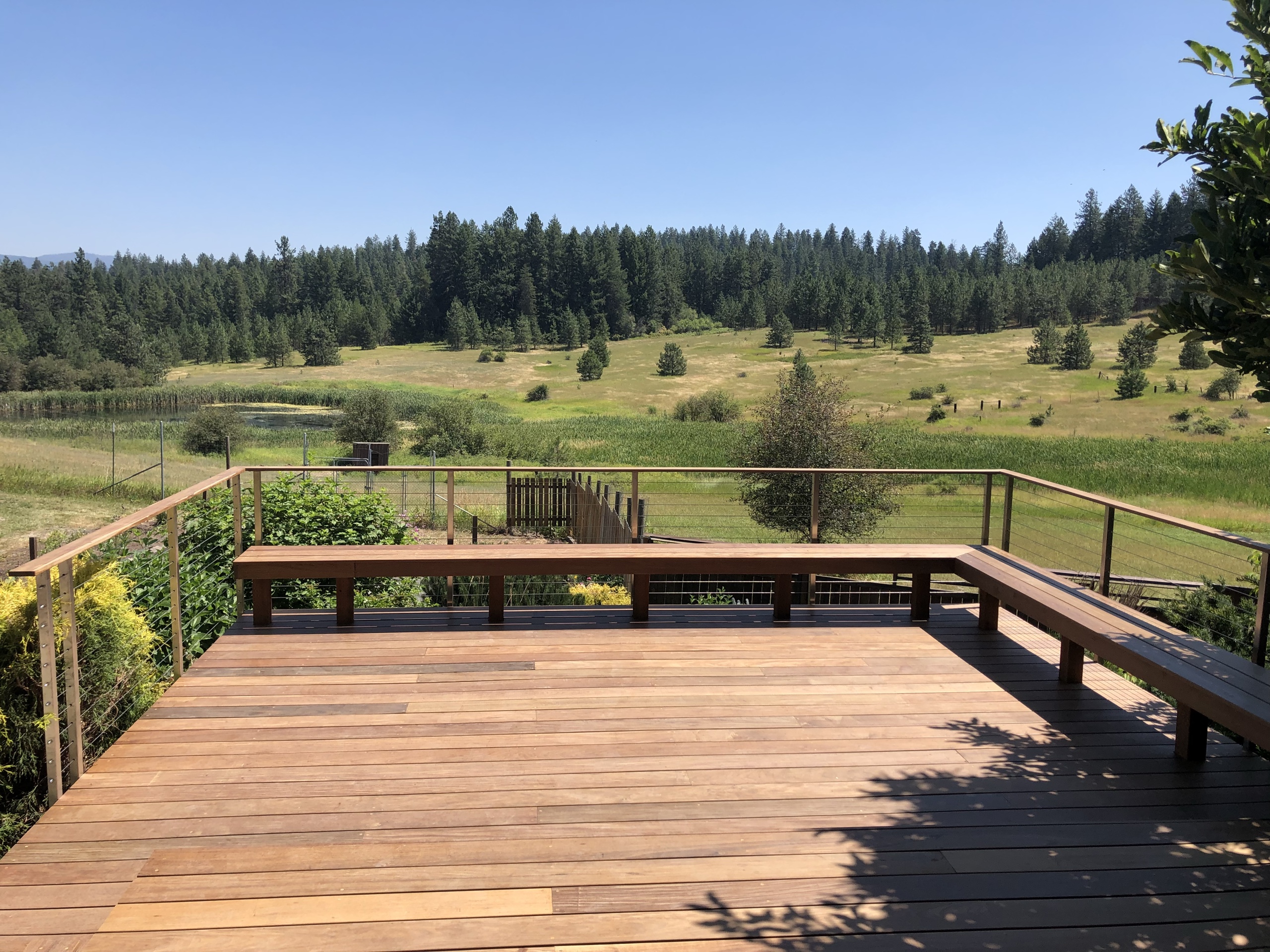 Large red-brown hardwood deck with built-in perimeter benches and cable railing facing a meadow