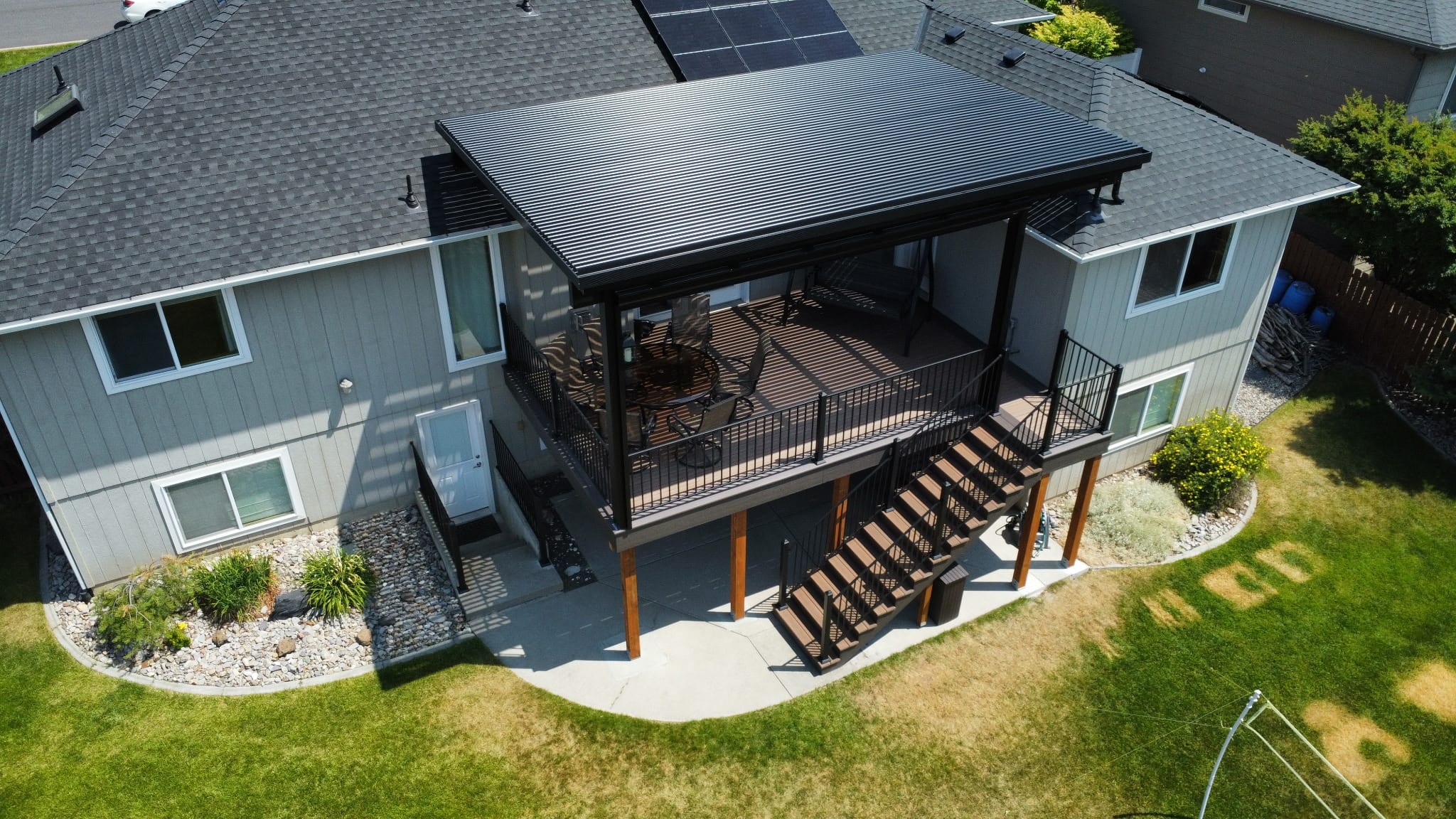 Aerial view of dark louvered pergola roof over an elevated composite deck with stairs, curved landscape beds and lawn