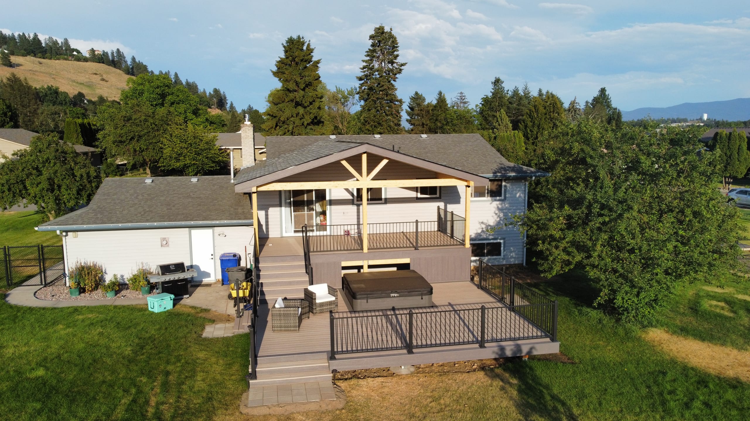 Aerial view at golden hour of house with gabled cedar-timber deck cover, hot tub and railed lower deck, hills behind