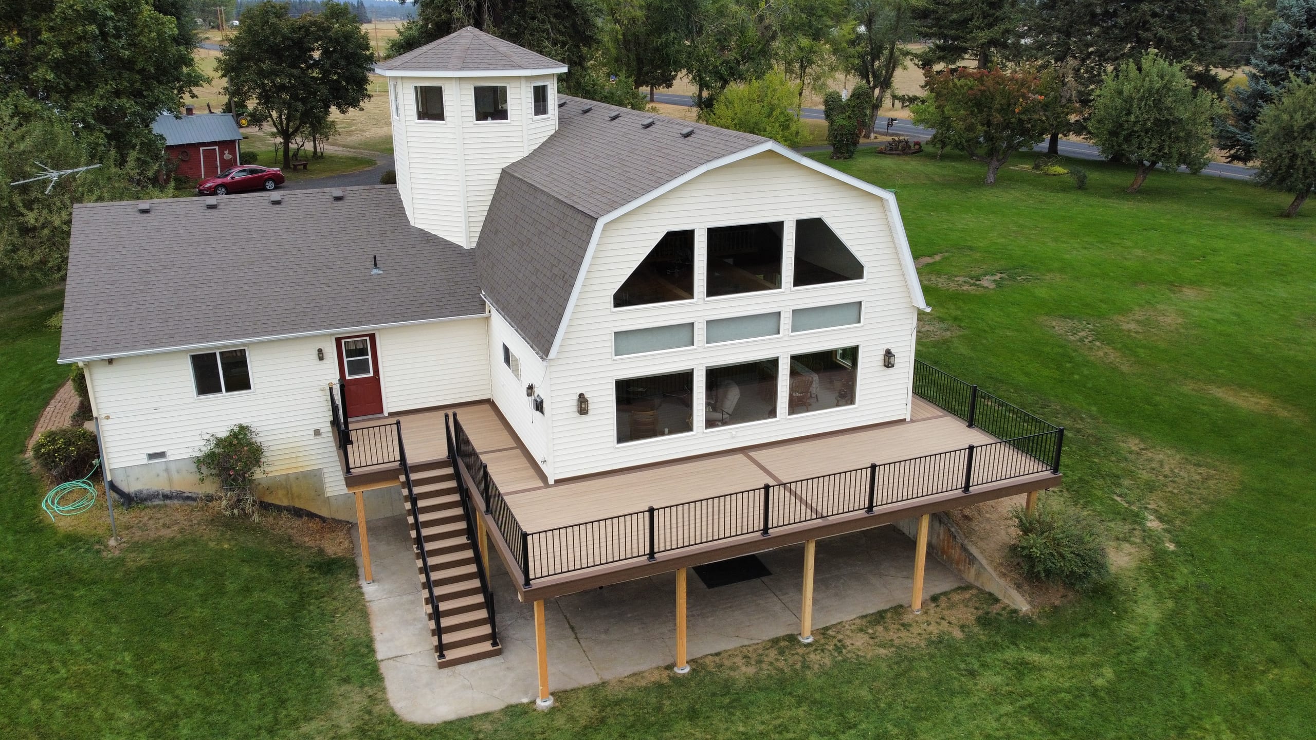 Aerial view of large tan composite deck with black railing and angled stairs at a white gambrel house in green fields