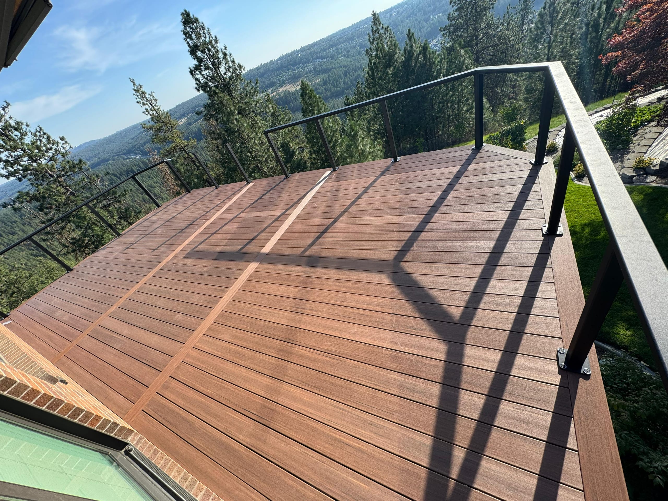 Aerial-style angle over mahogany-toned composite deck with diagonal borders and black metal railing above a forested valley