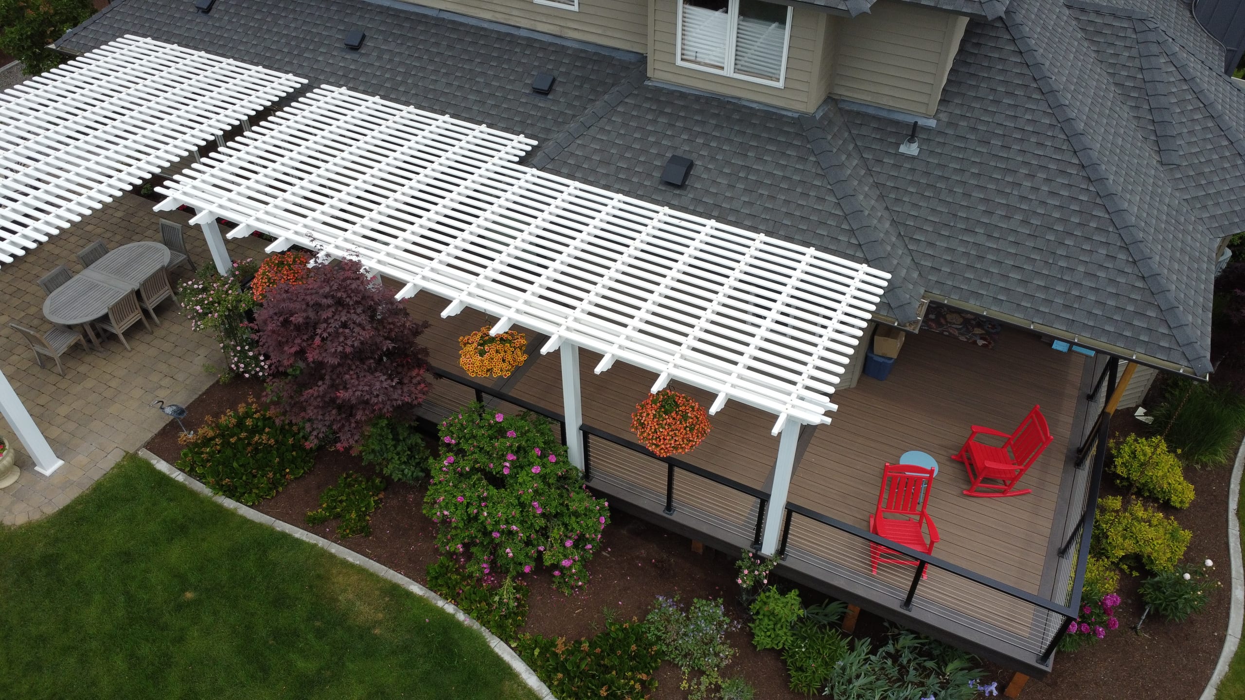 Aerial view of white lattice pergola over backyard deck with red chairs, patio table, plantings beside gray-roofed house
