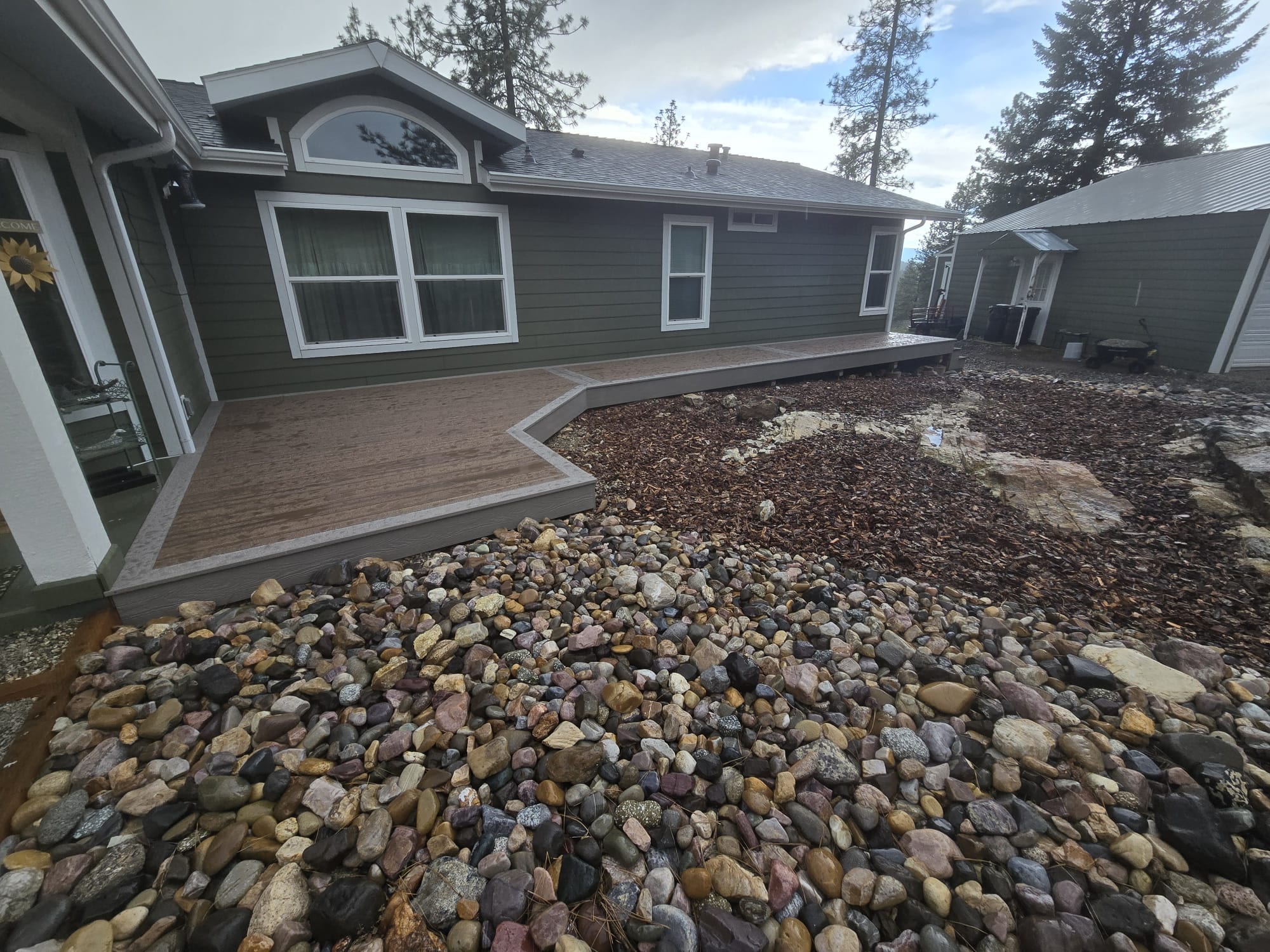Angled brown composite walkway deck with white trim beside a green house, river-rock landscaping