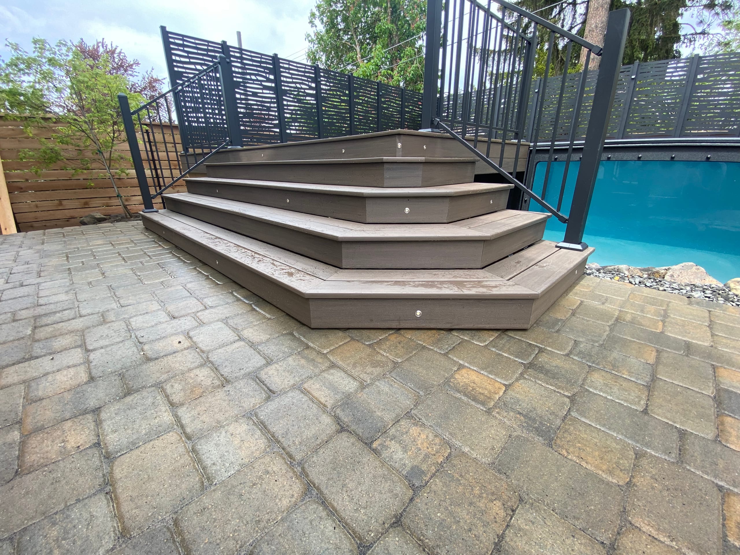 Angled composite steps with step lights down to paver patio beside a raised pool with privacy screen