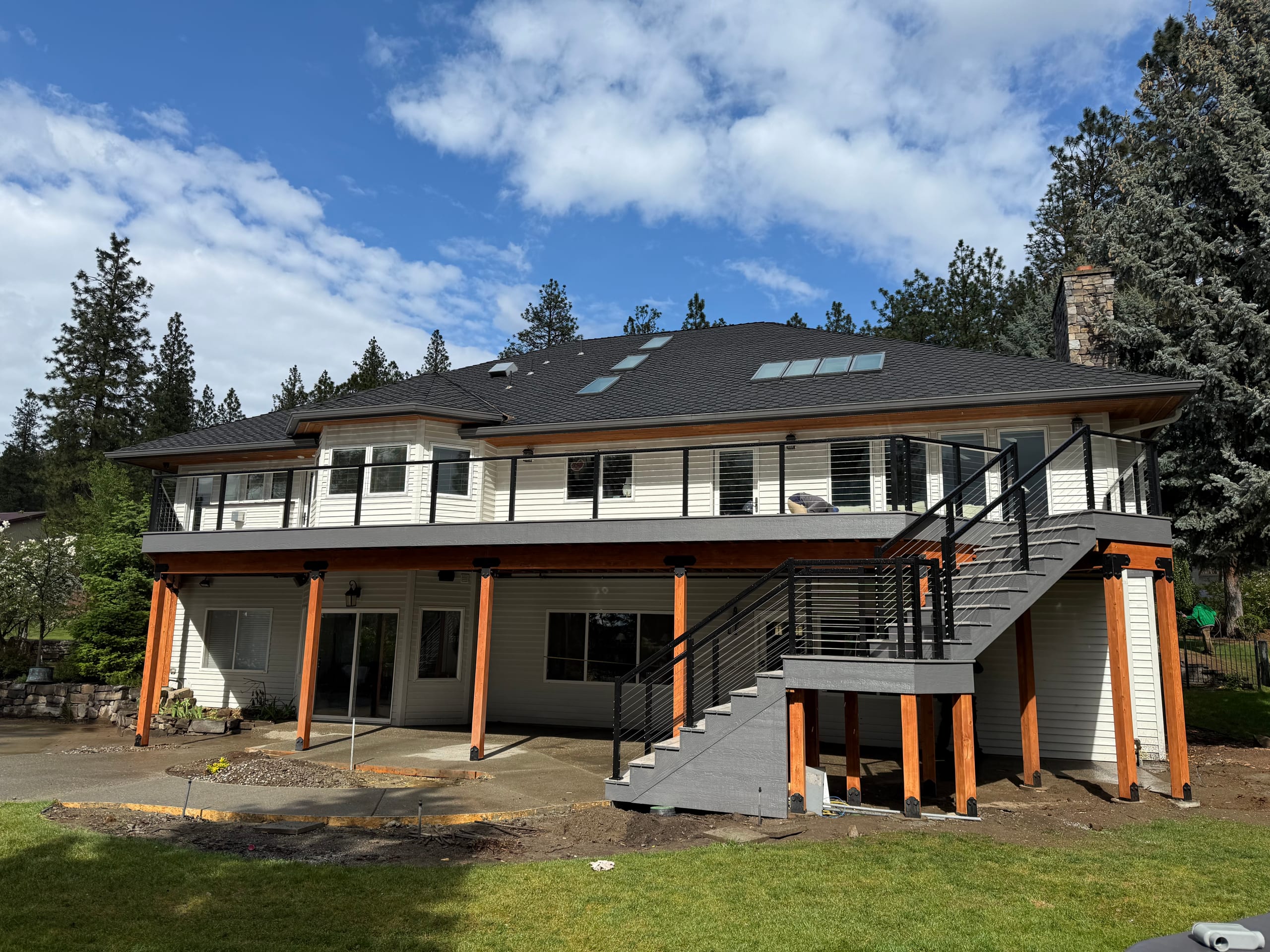 Back of large two-story home with elevated wrap deck on cedar-wrapped posts, black railing and metal stairs, blue sky