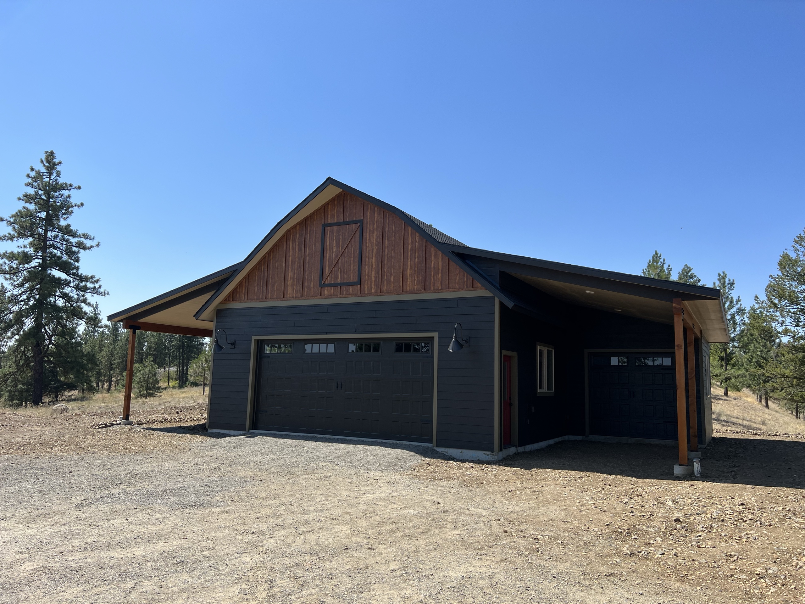 Dark barn-style garage with a cedar gable and a lean-to carport on timber posts, pines behind