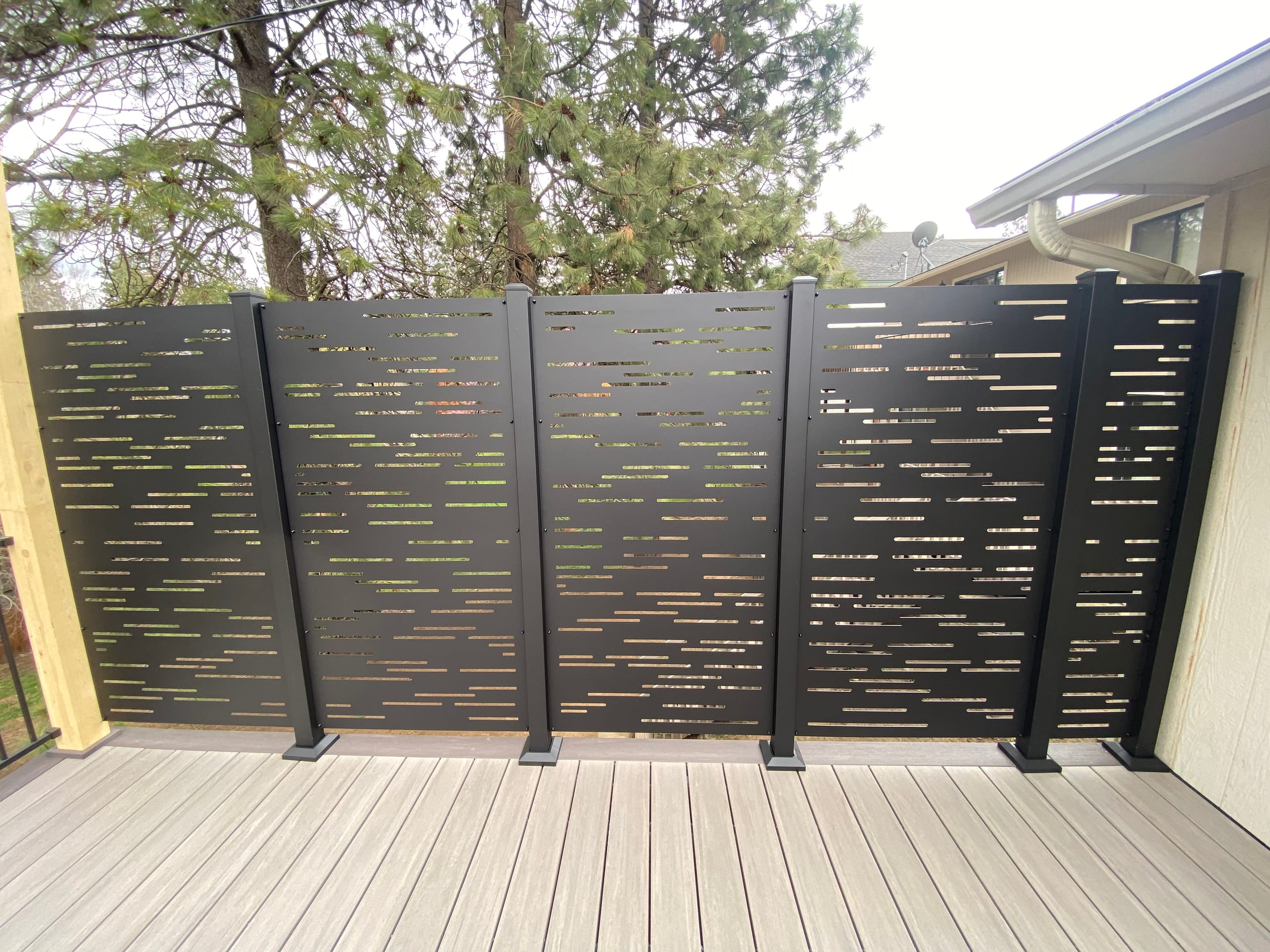 Black perforated metal privacy screen panels on gray composite deck beside a house, pine trees behind