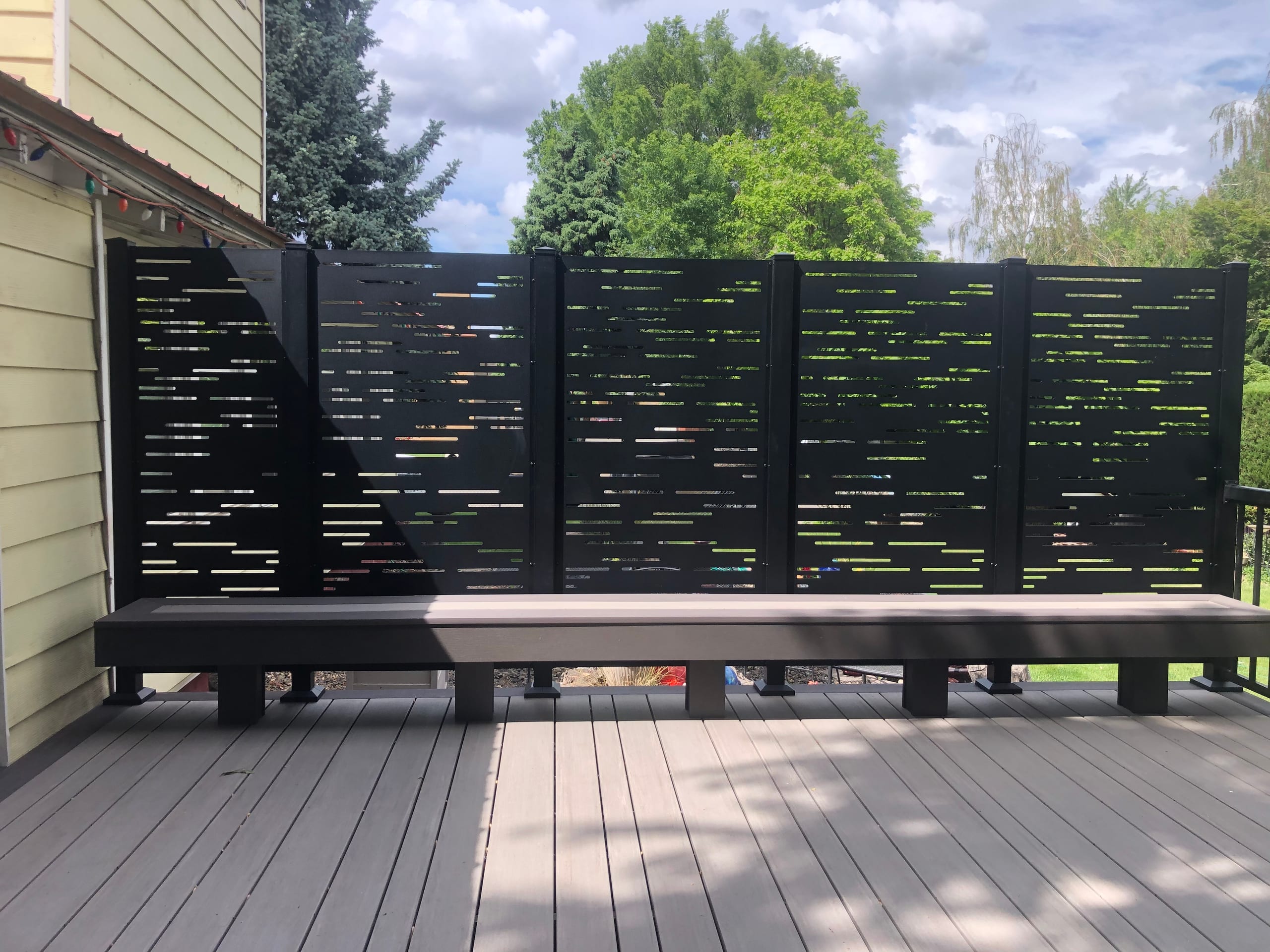 Black slatted privacy screen above built-in bench on light gray composite deck, trees behind