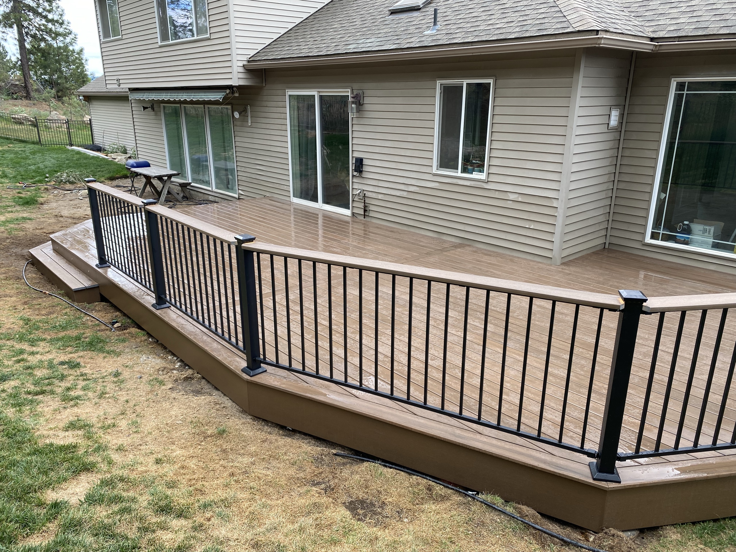 Brown composite deck with angled black picket railing along back of tan house, dry lawn