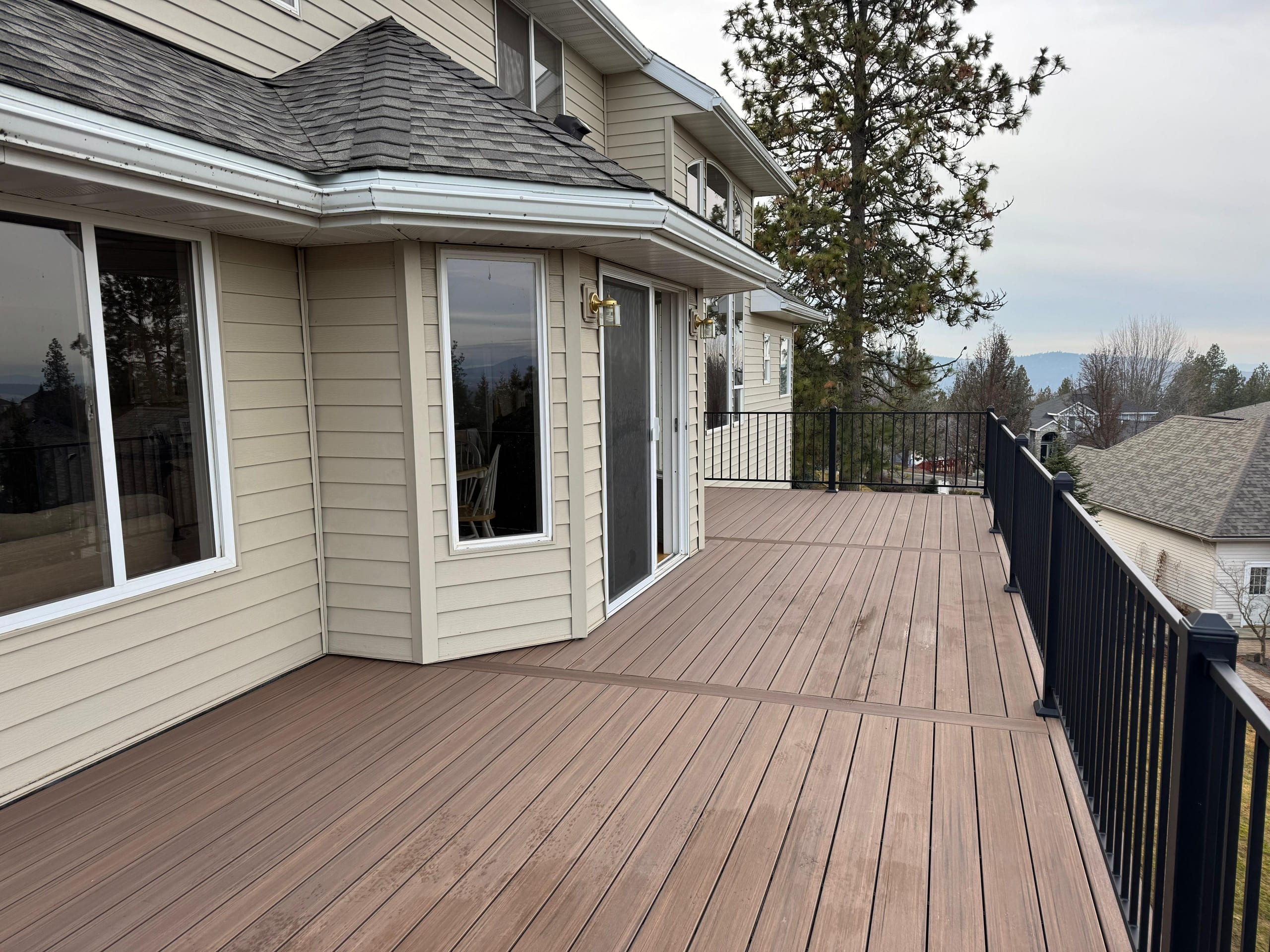 Brown composite deck along beige house with black picket railing and bay windows, hillside neighborhood beyond