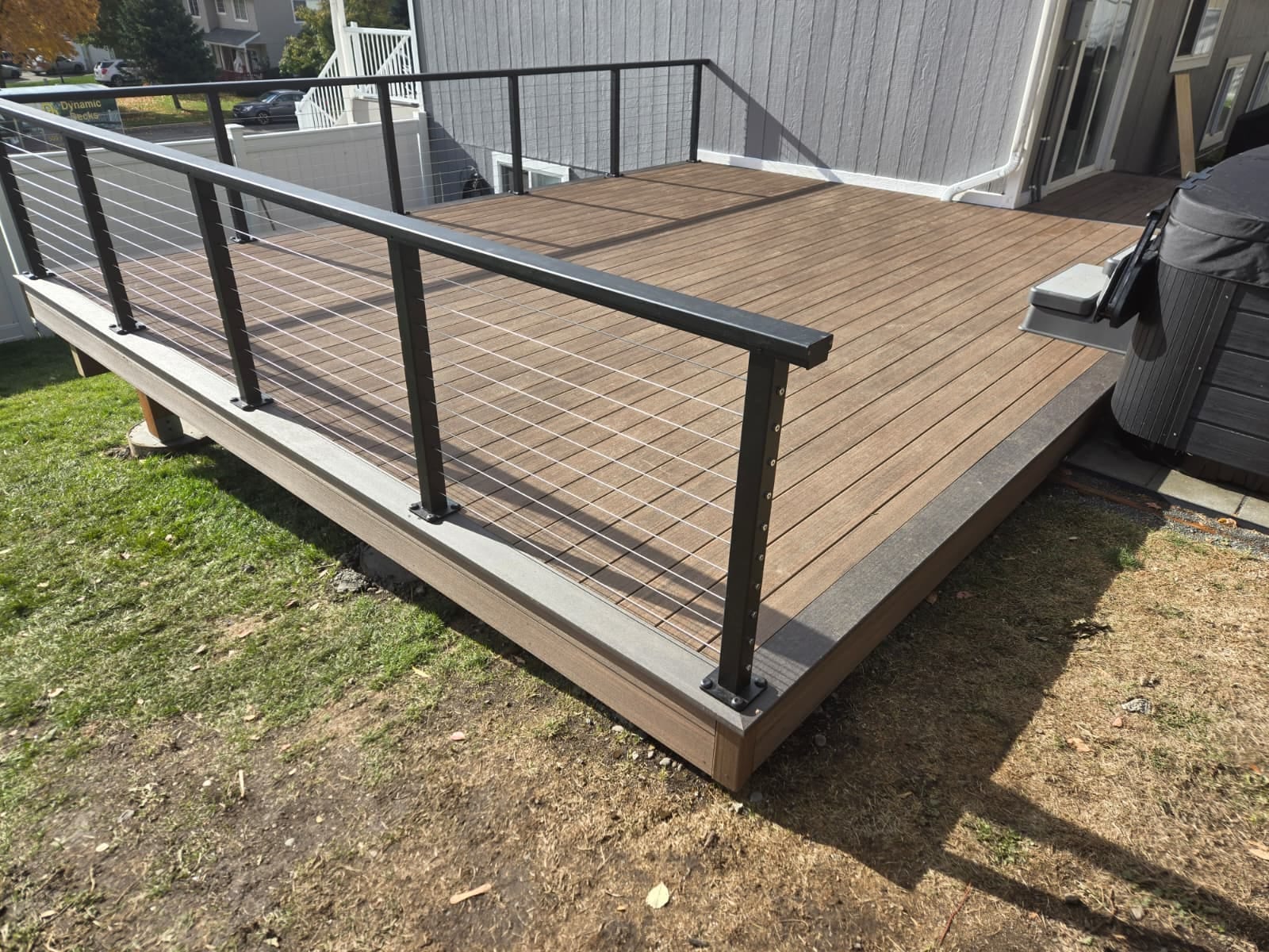Brown composite deck with black cable railing and tan fascia in a lawn beside a house, hot tub at right