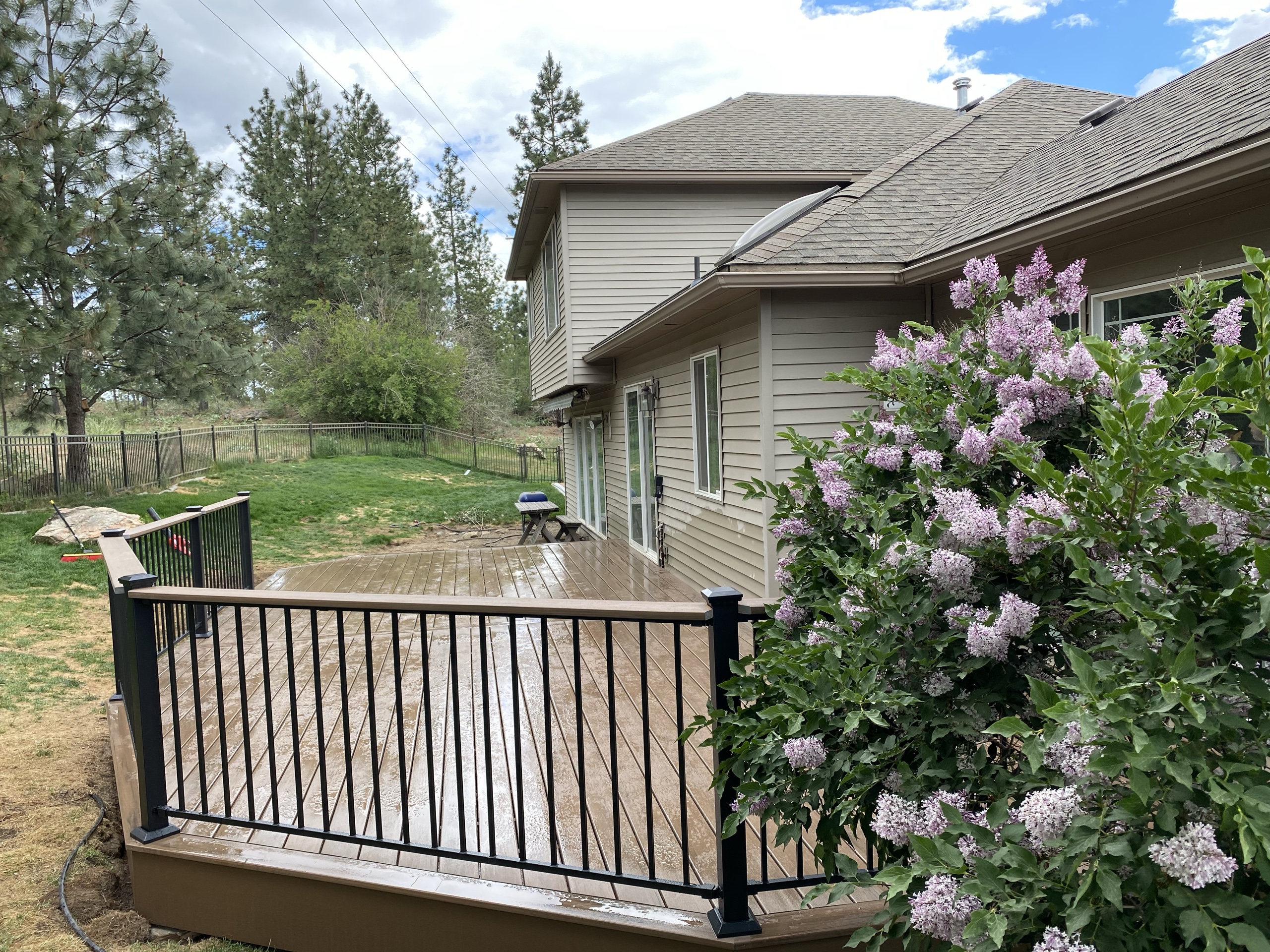 Brown composite deck with black picket railing behind lilac bush, tan house and trees