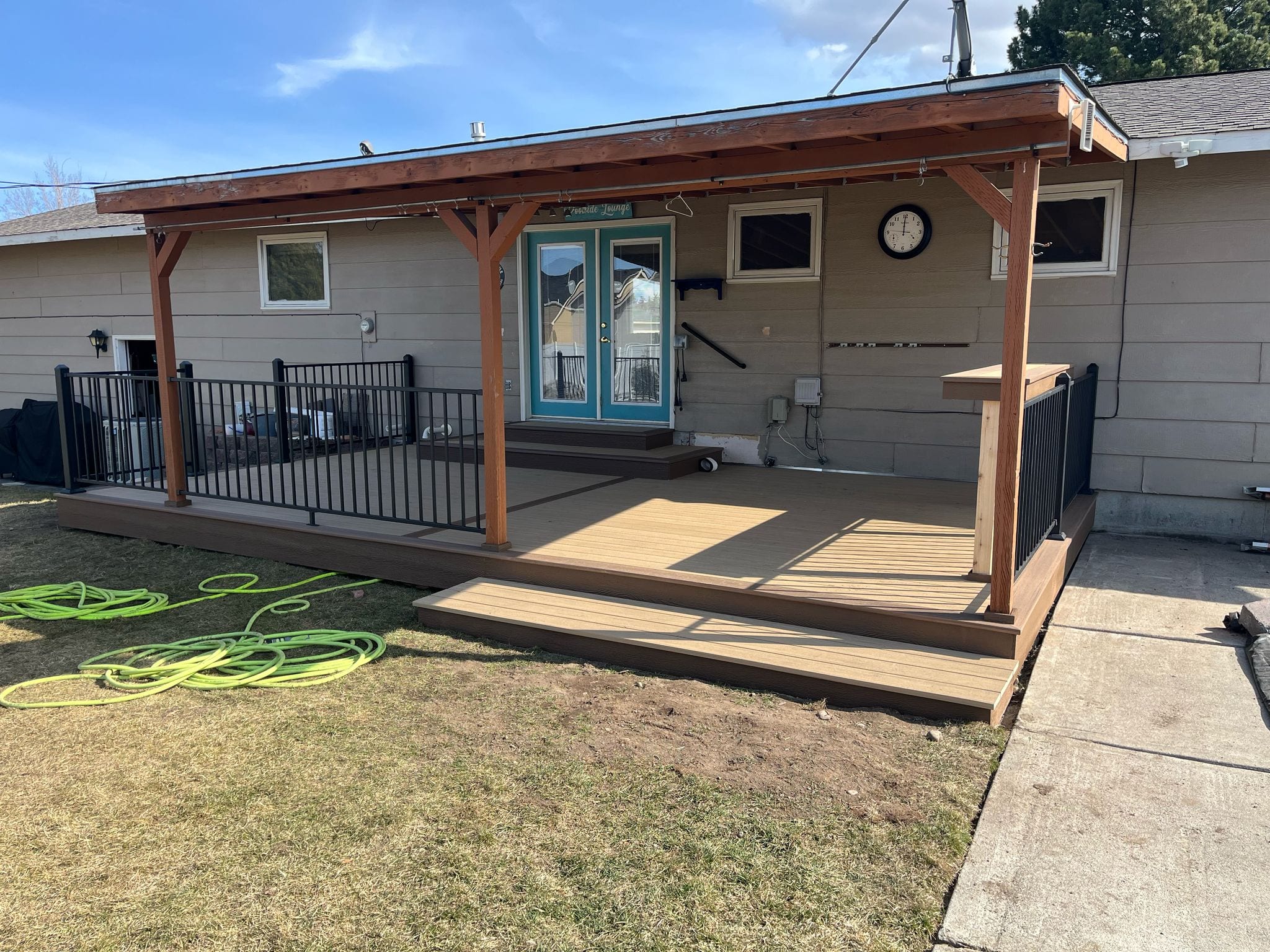 Brown composite deck with cedar-post patio cover, black metal railing and bar counter at a rambler, lawn with hoses