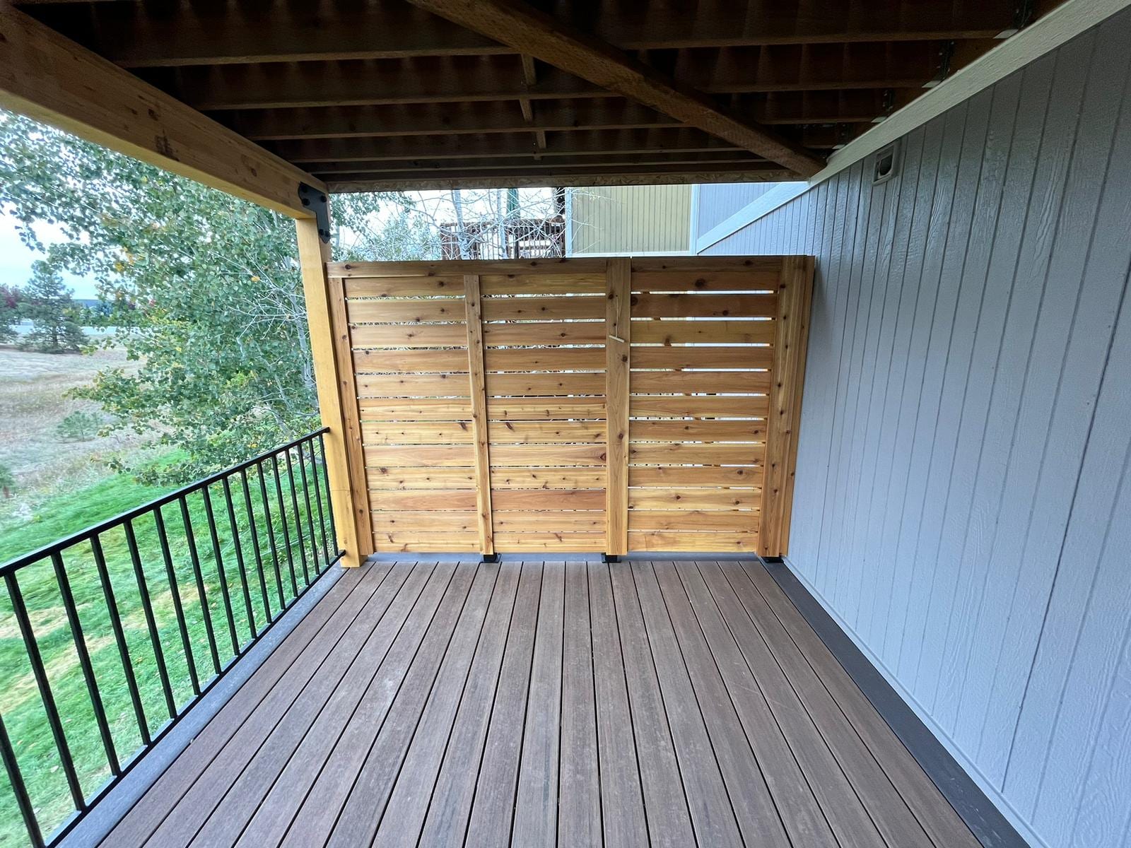 Brown composite deck under a covered upper deck with cedar horizontal privacy wall and black metal picket railing