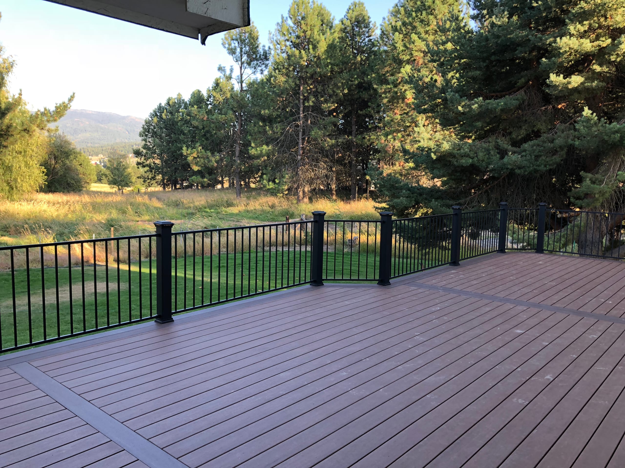 Brown composite deck with dark border and black picket railing with post caps facing a green meadow and pines