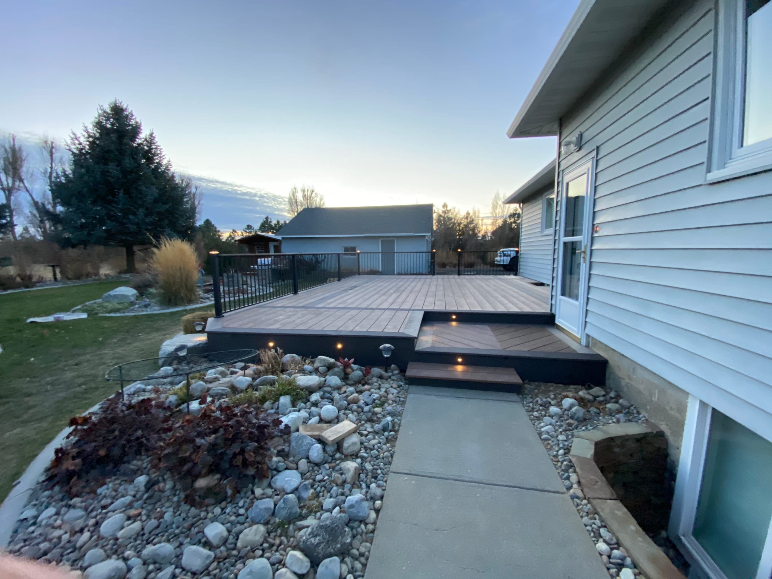 Brown composite deck with lit step and black railing beside pale house and concrete walkway through rock garden at sunset