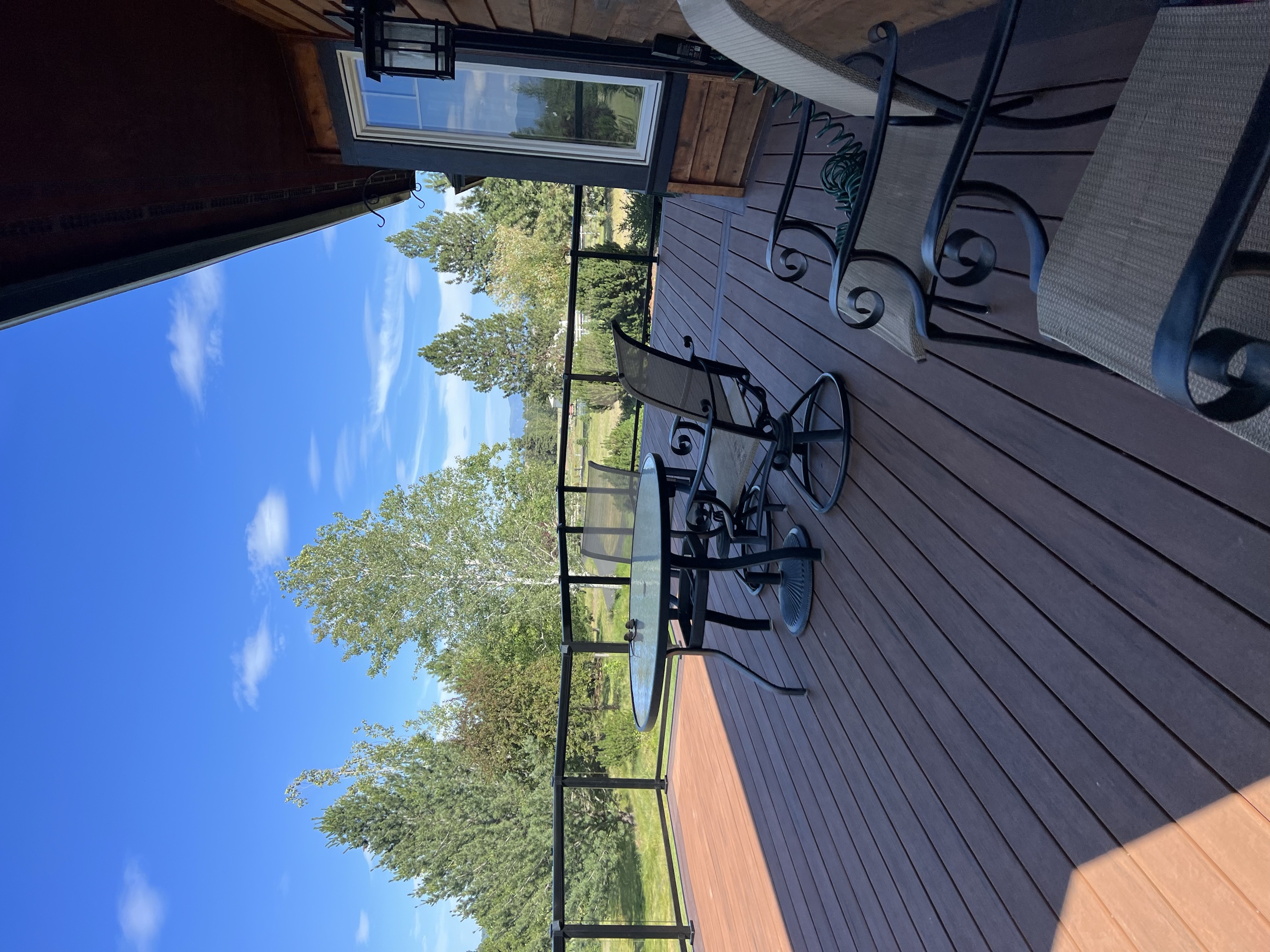 Brown composite deck with patio table and chairs, black railing, cedar house wall and trees
