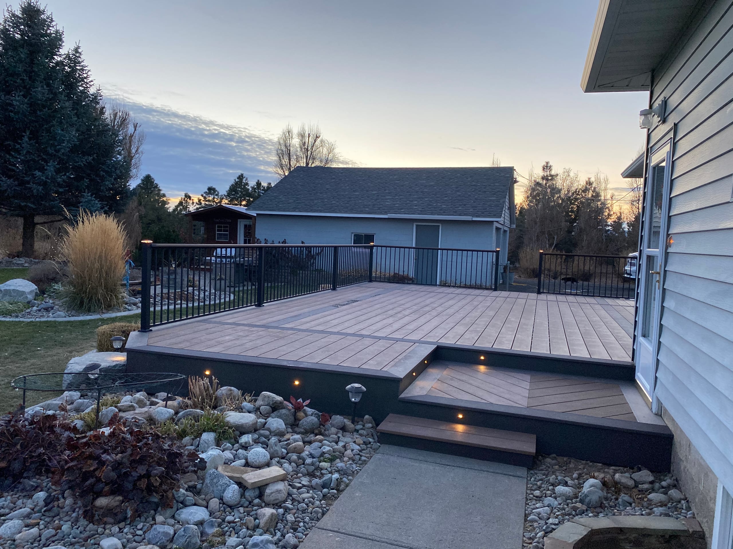 Brown composite deck with picture-frame border and lit steps beside house at sunset, black railing, rock landscaping
