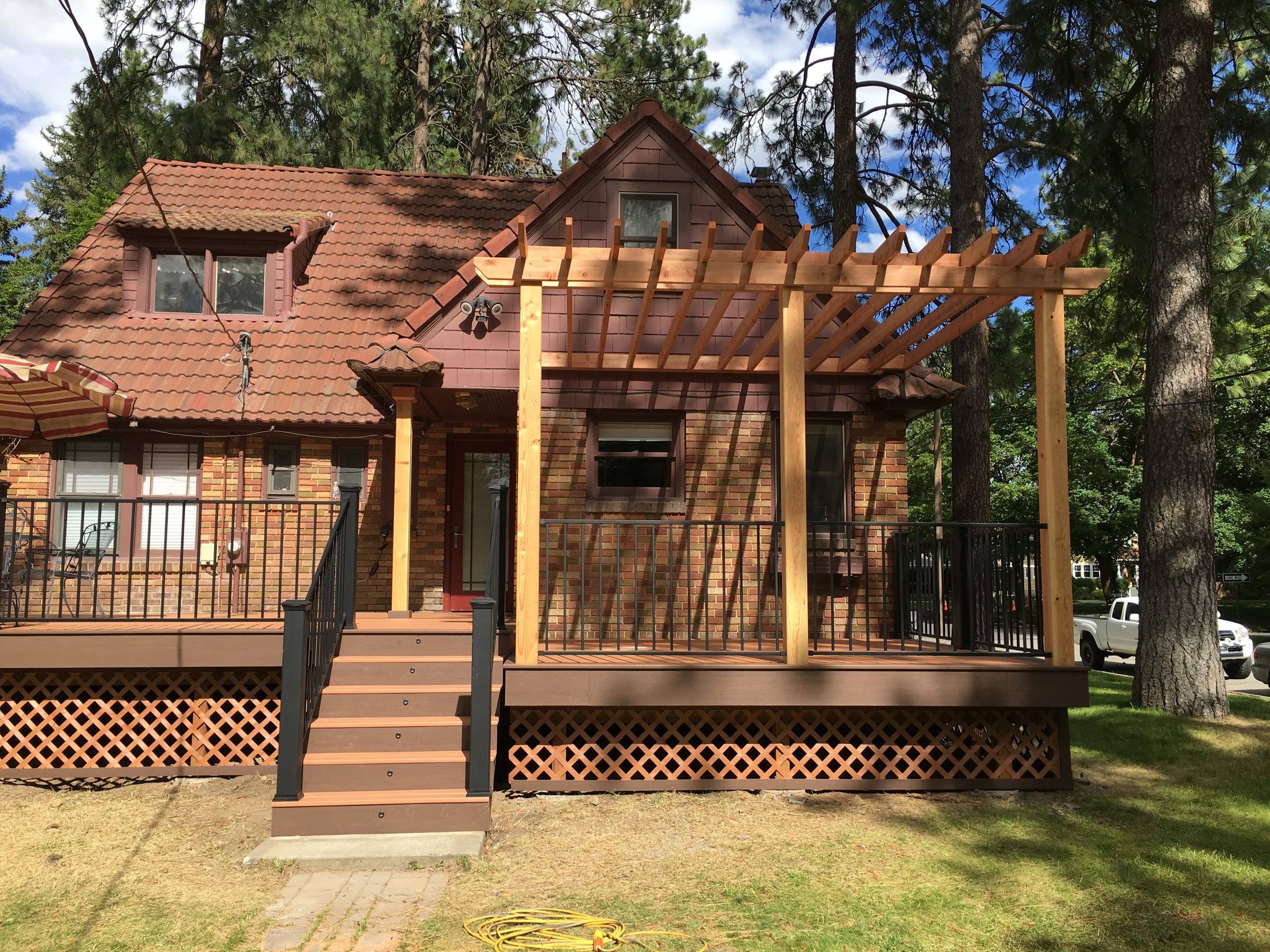 Brown deck with lattice skirt, wood pergola and black railing and steps at a brick-and-shingle home