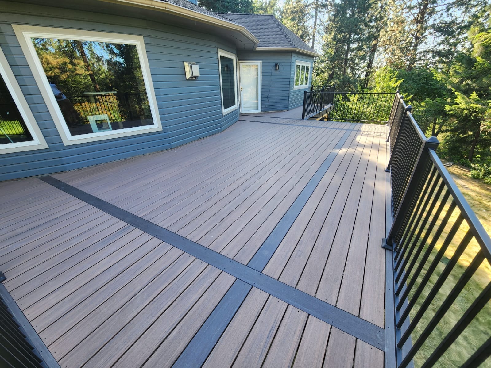 Brown-gray composite deck with dark diagonal inlay stripes and black metal picket railing along blue house, pines