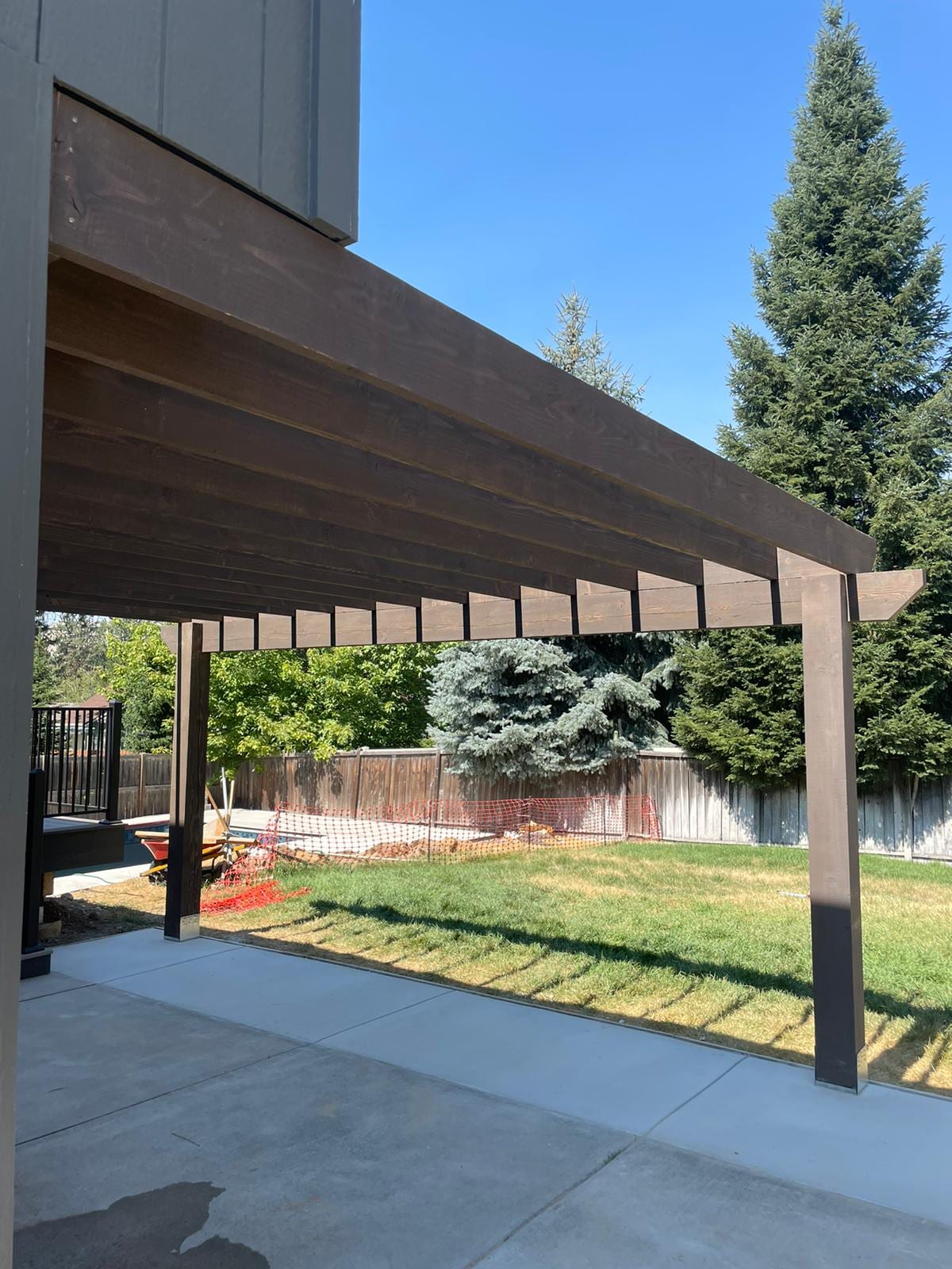 Brown pergola beam and slat structure attached to house, over a new concrete patio with lawn and trees