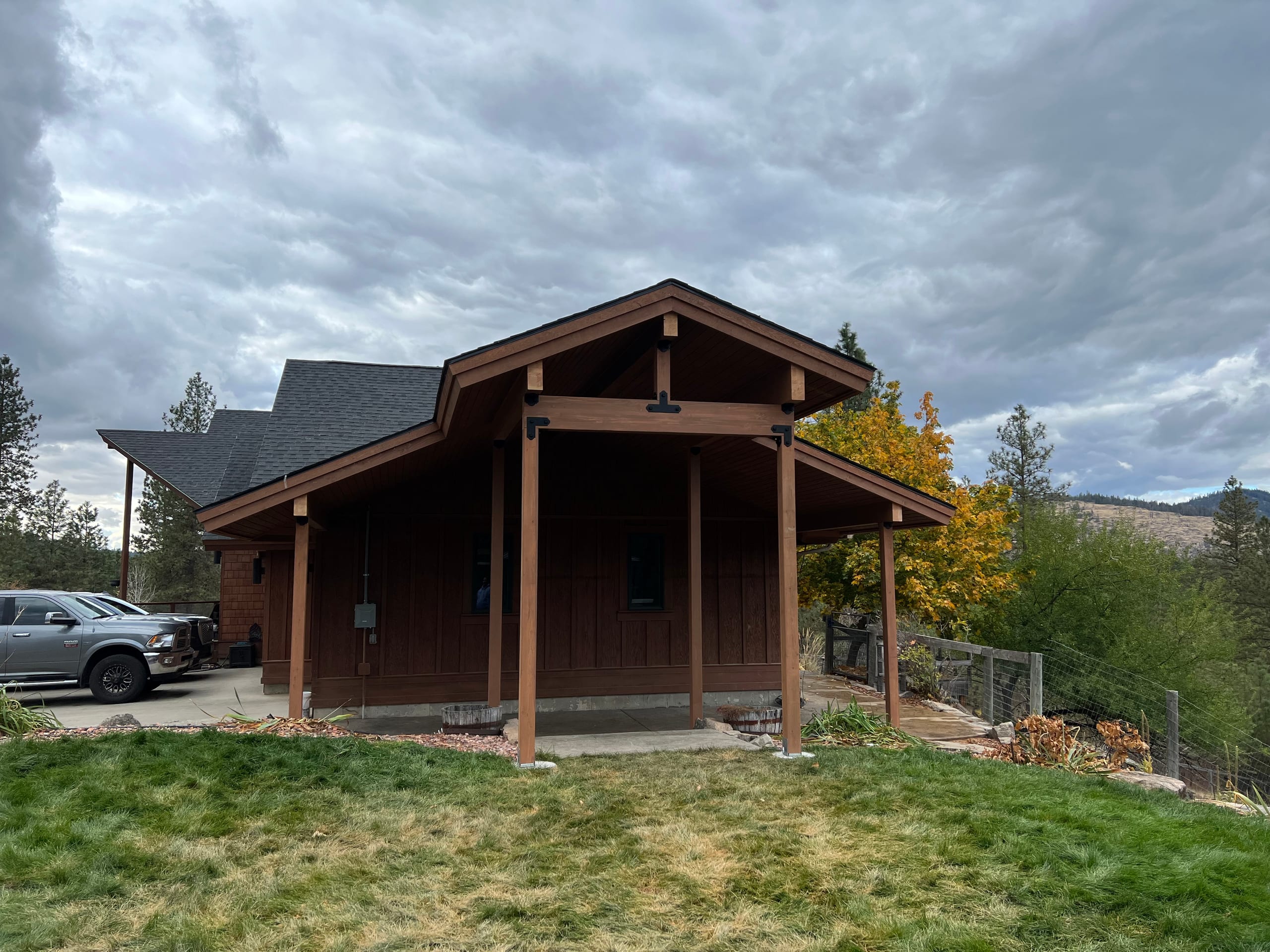 Brown timber-frame gabled covered porch/carport extension with pickup truck, autumn tree and hills, cloudy sky
