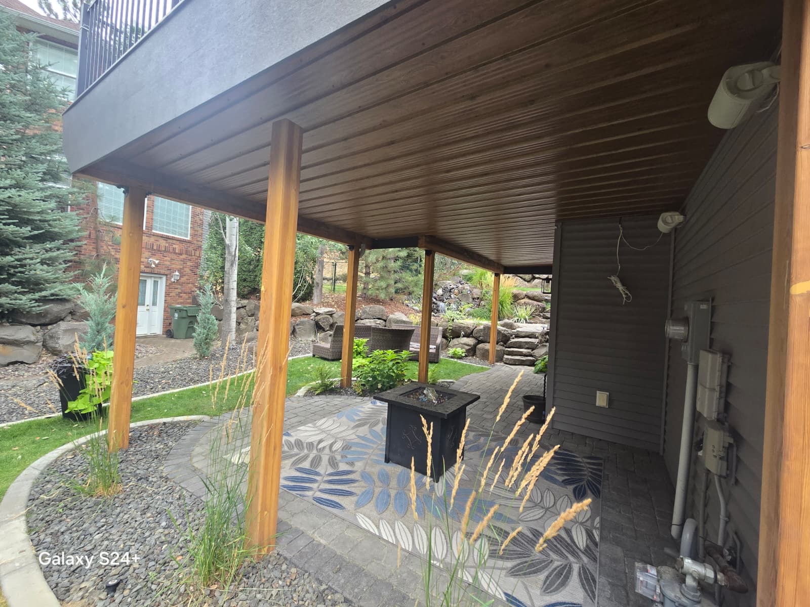 Brown underdeck ceiling with cedar posts over a patio with fire table, landscaped garden and boulders beyond