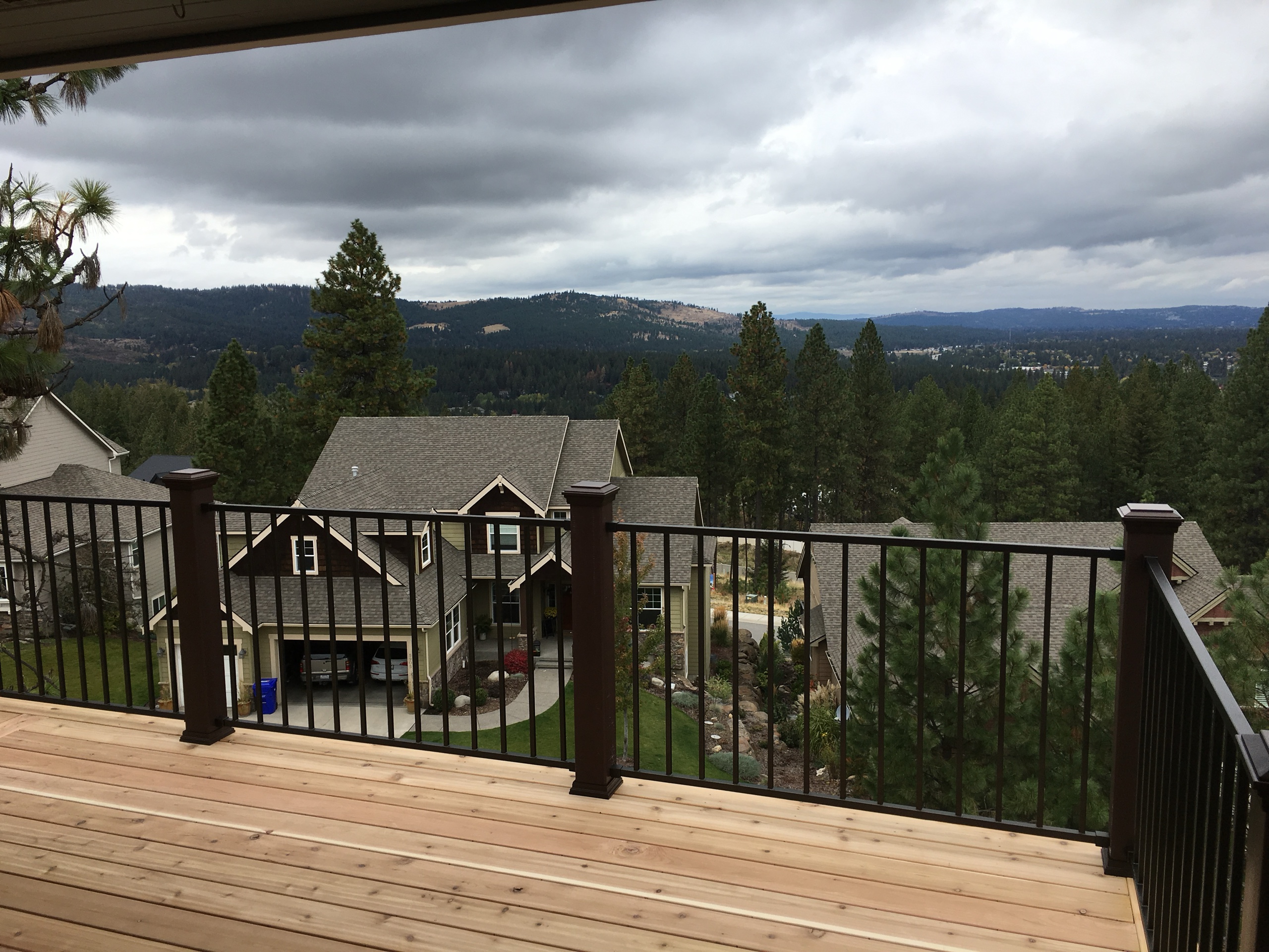 View from cedar deck over black picket railing toward wooded valley and neighboring house, stormy sky