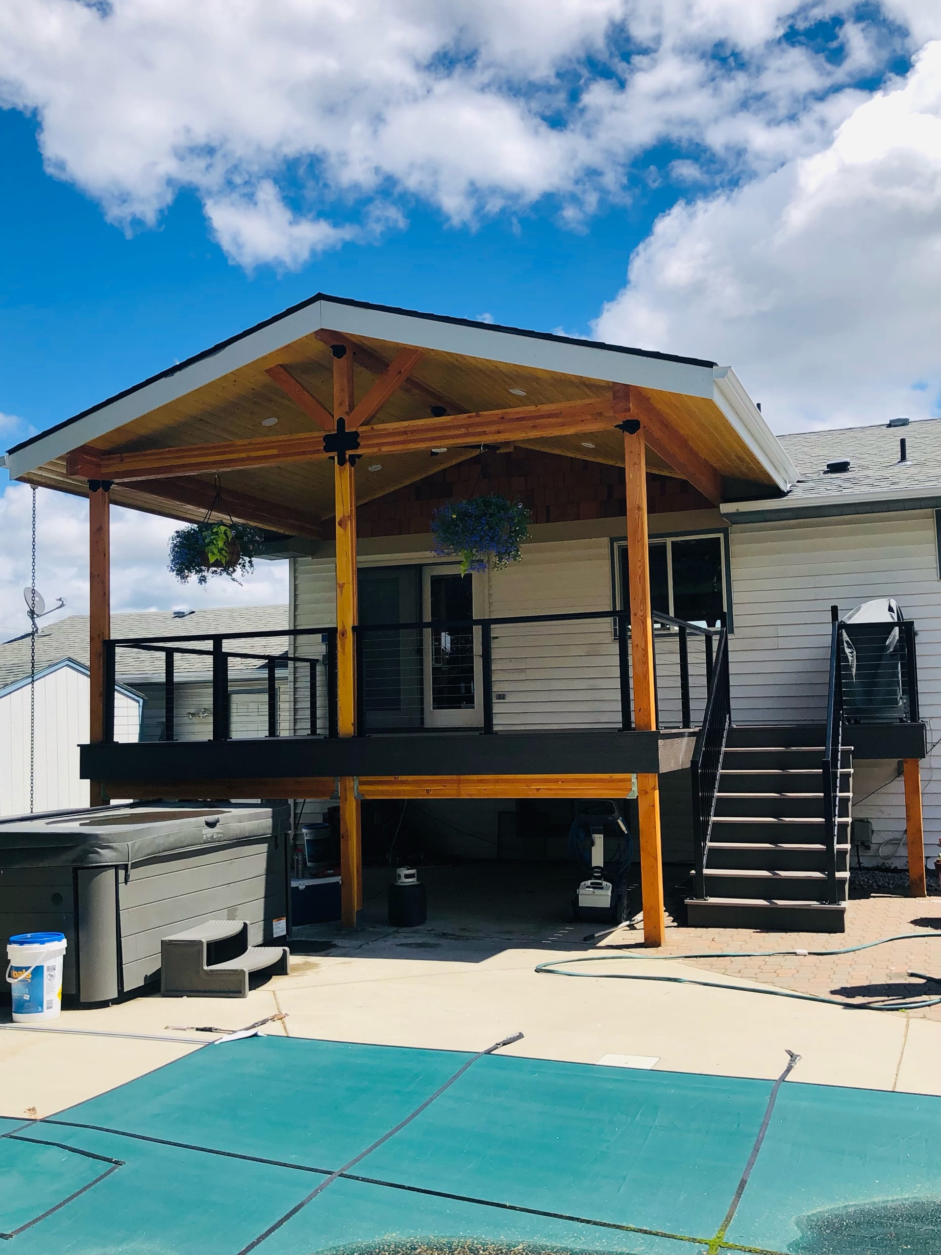 Cedar gabled covered deck on stilts with black railing and stair above a pool with teal safety cover and hot tub, blue sky