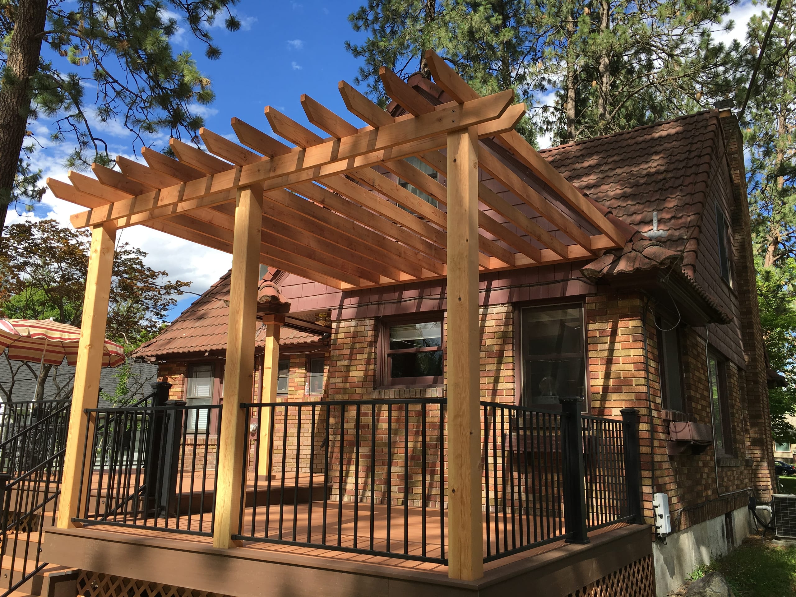Cedar pergola over a wood deck with black railing at a shingled house, trees behind