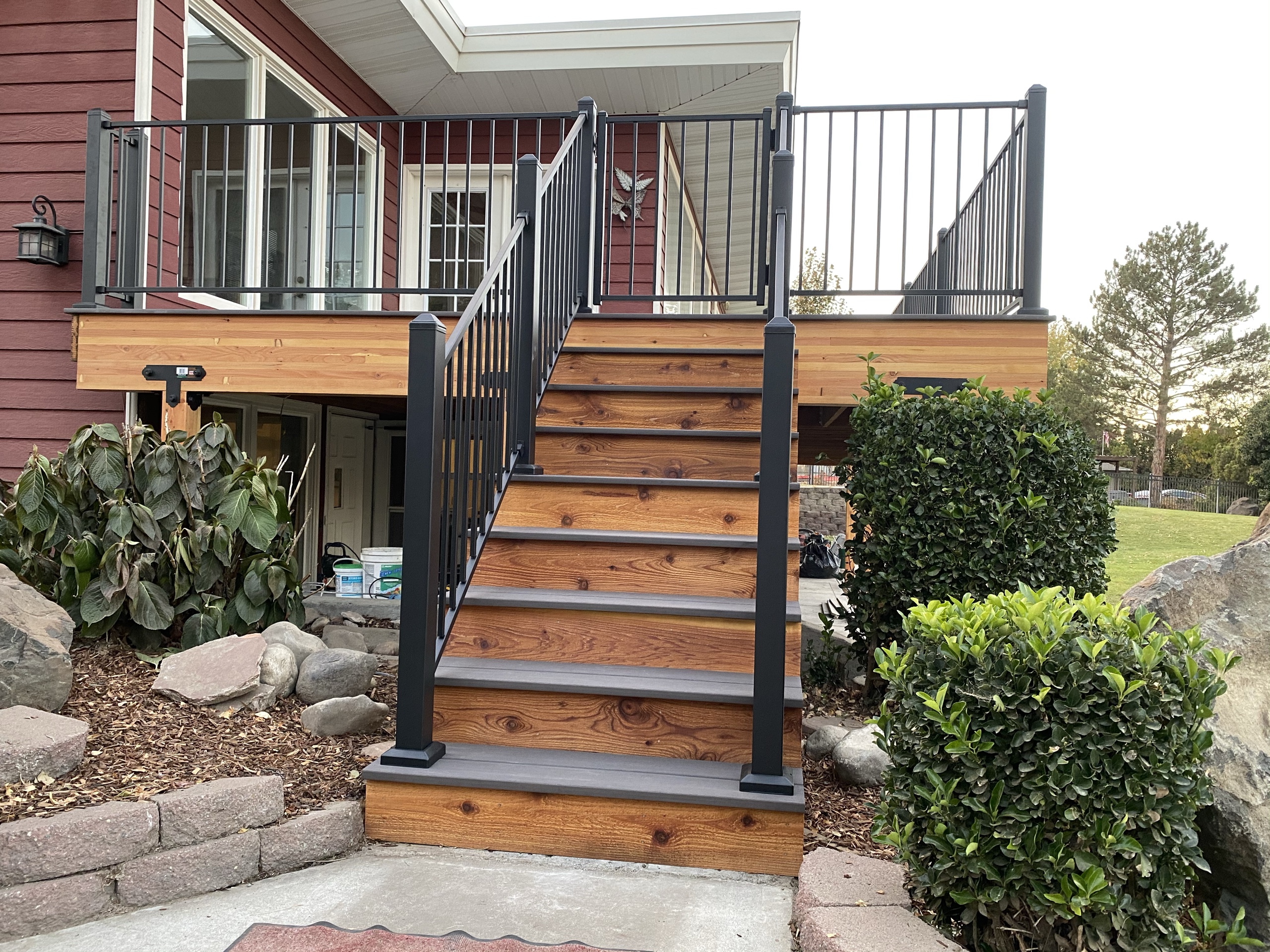 Cedar stair with gray treads and black railing leading to elevated deck on red house, rock landscaping