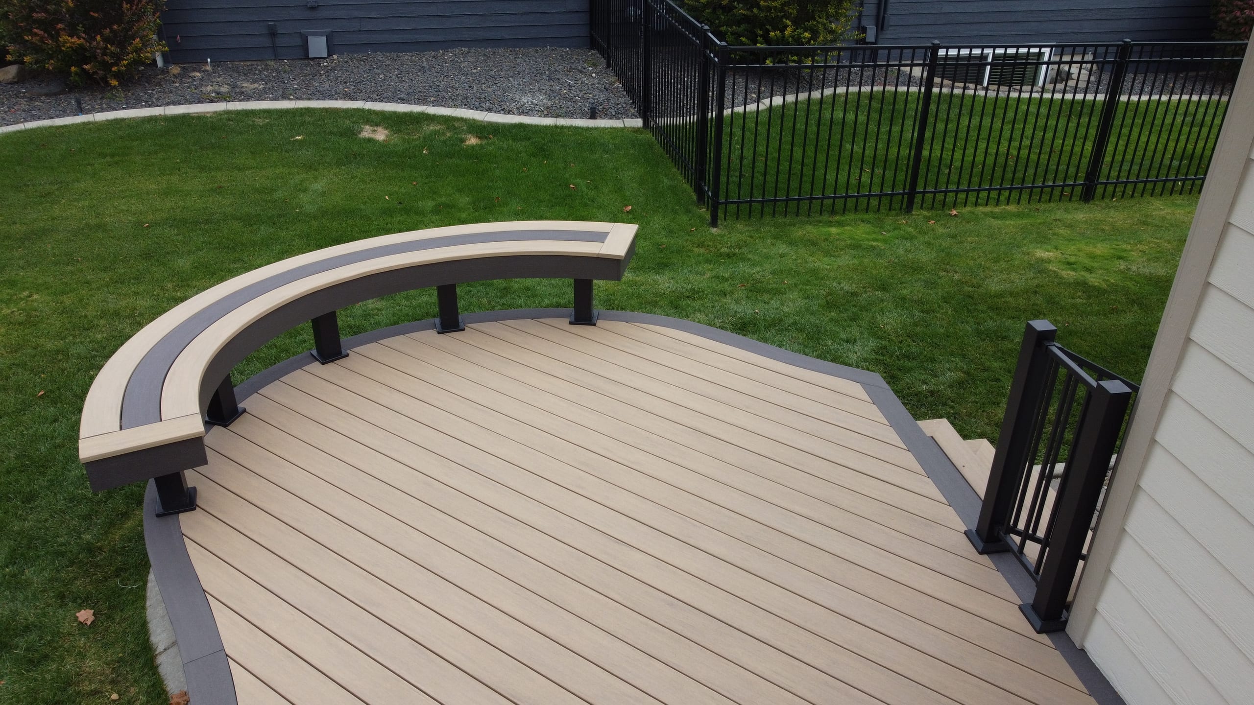 Close view of curved bench on beige composite deck with gray border, lawn and black fence