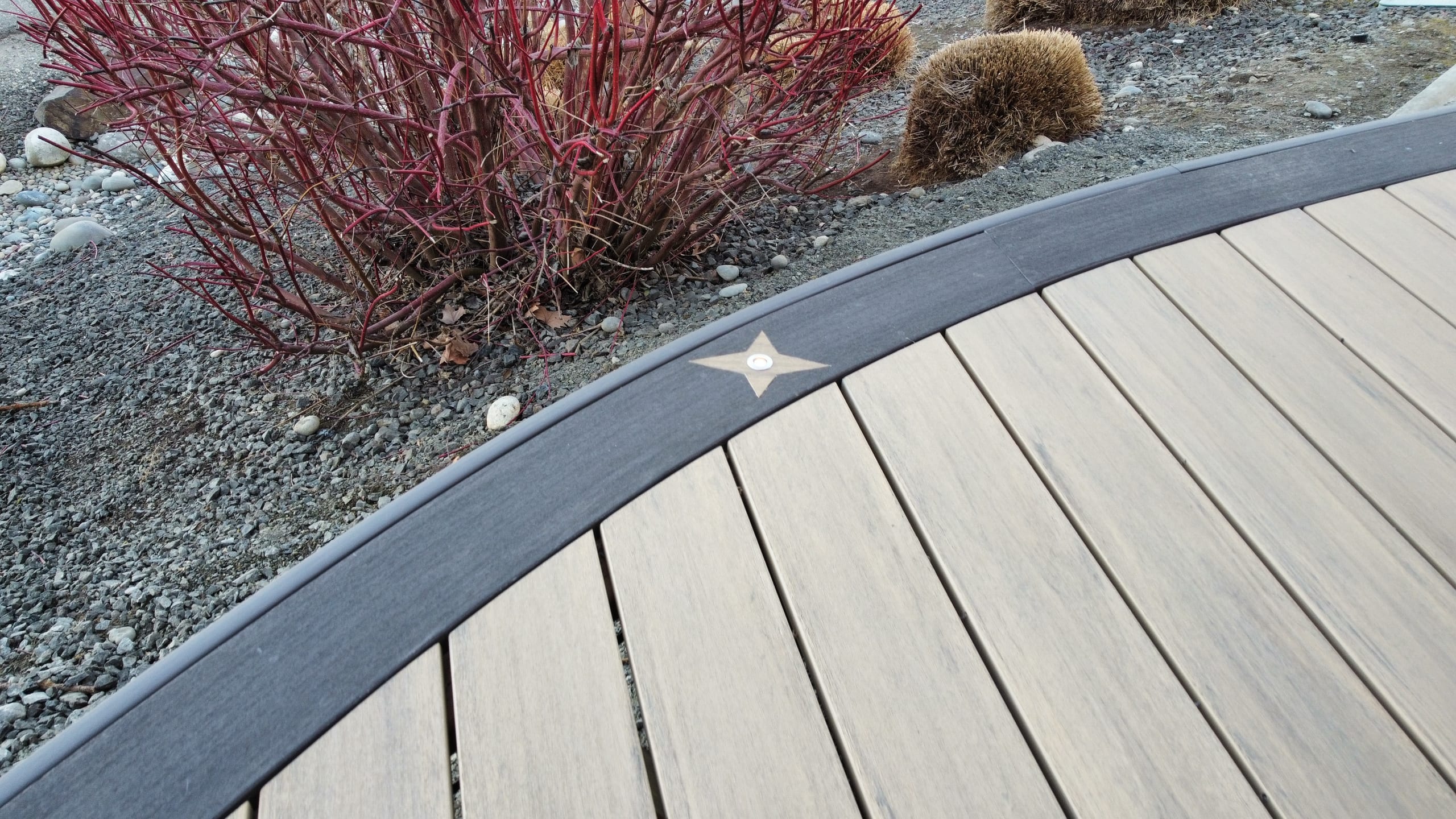 Close view of curved composite deck edge with dark border and star-shaped inlay accent, gravel and red-twig shrub