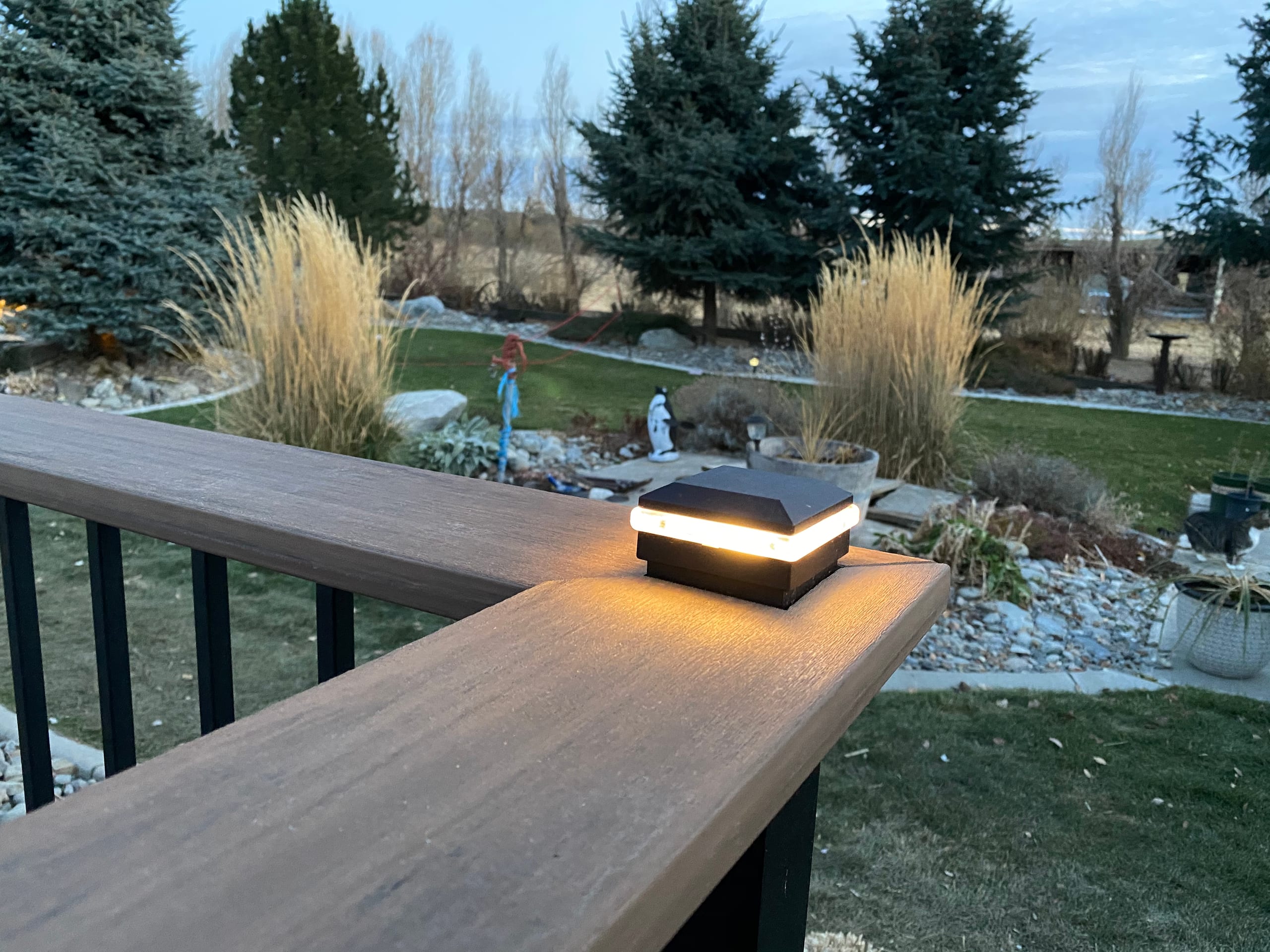 Close-up of composite railing cap with glowing lantern-style post light, rock garden and evergreens behind