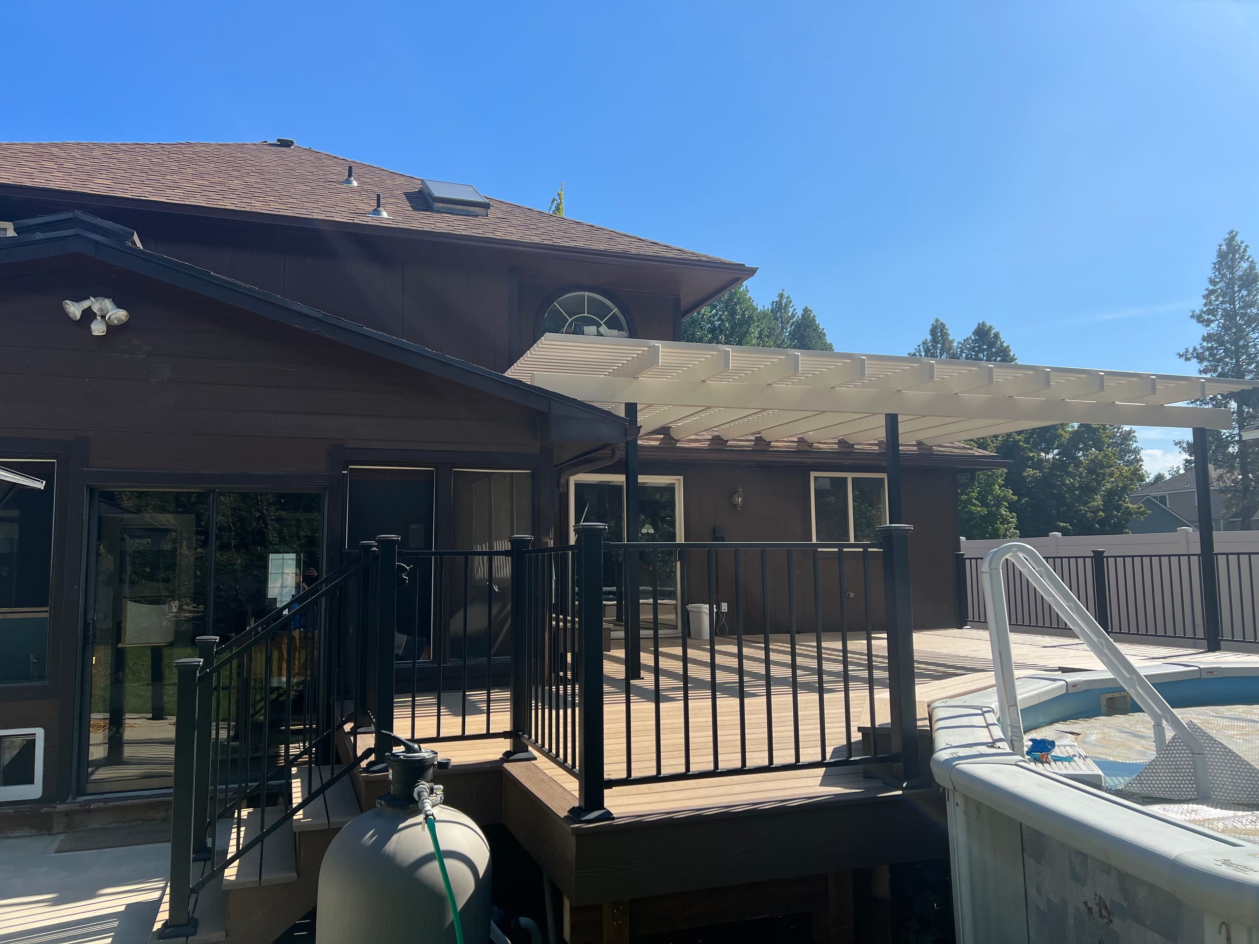 Composite pool deck with black metal railing and cream pergola/patio cover against dark brown house, pool cover in foreground