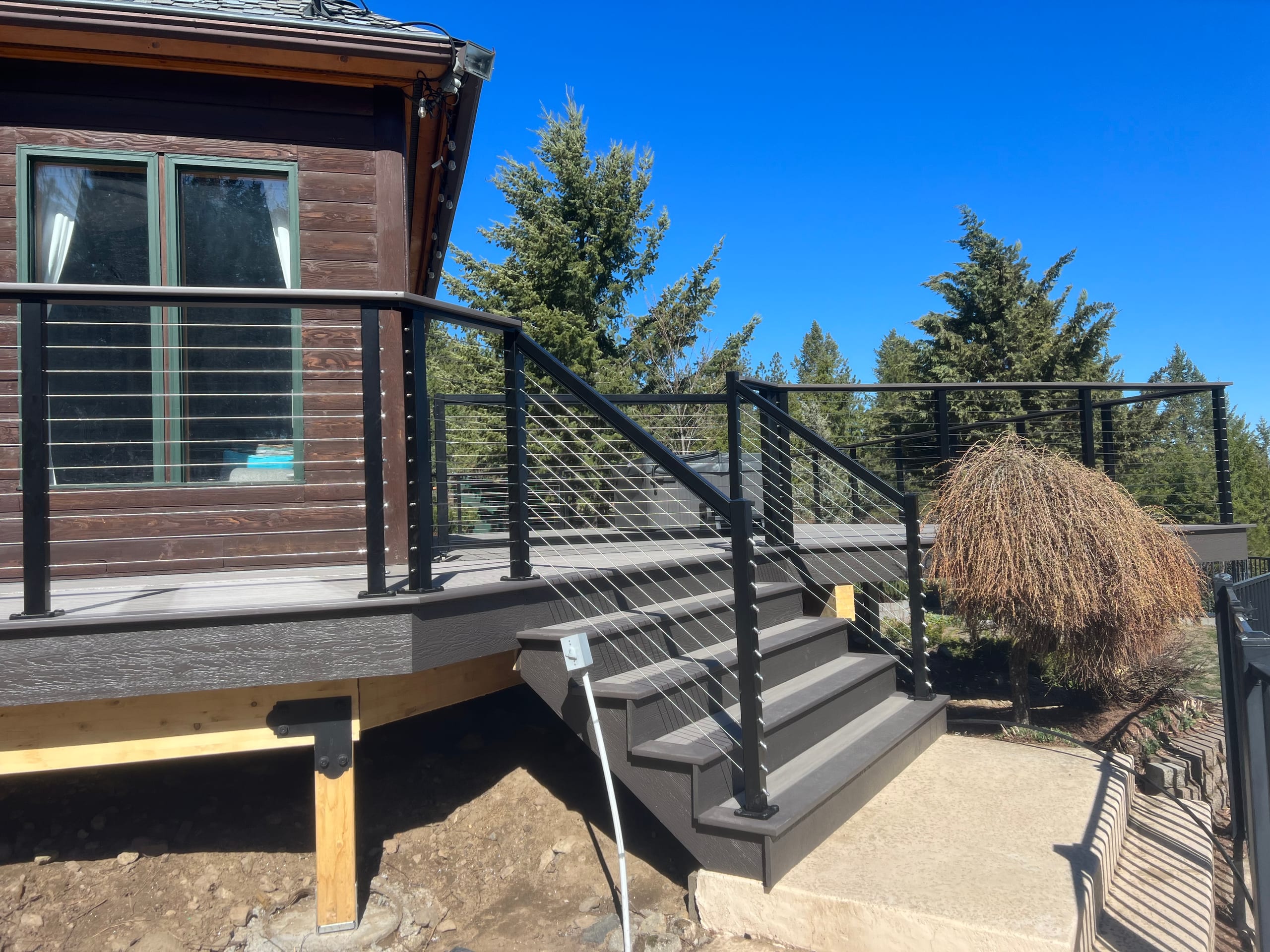 Composite stairs with black cable railing leading down from an elevated deck beside a cedar-stained house, blue sky