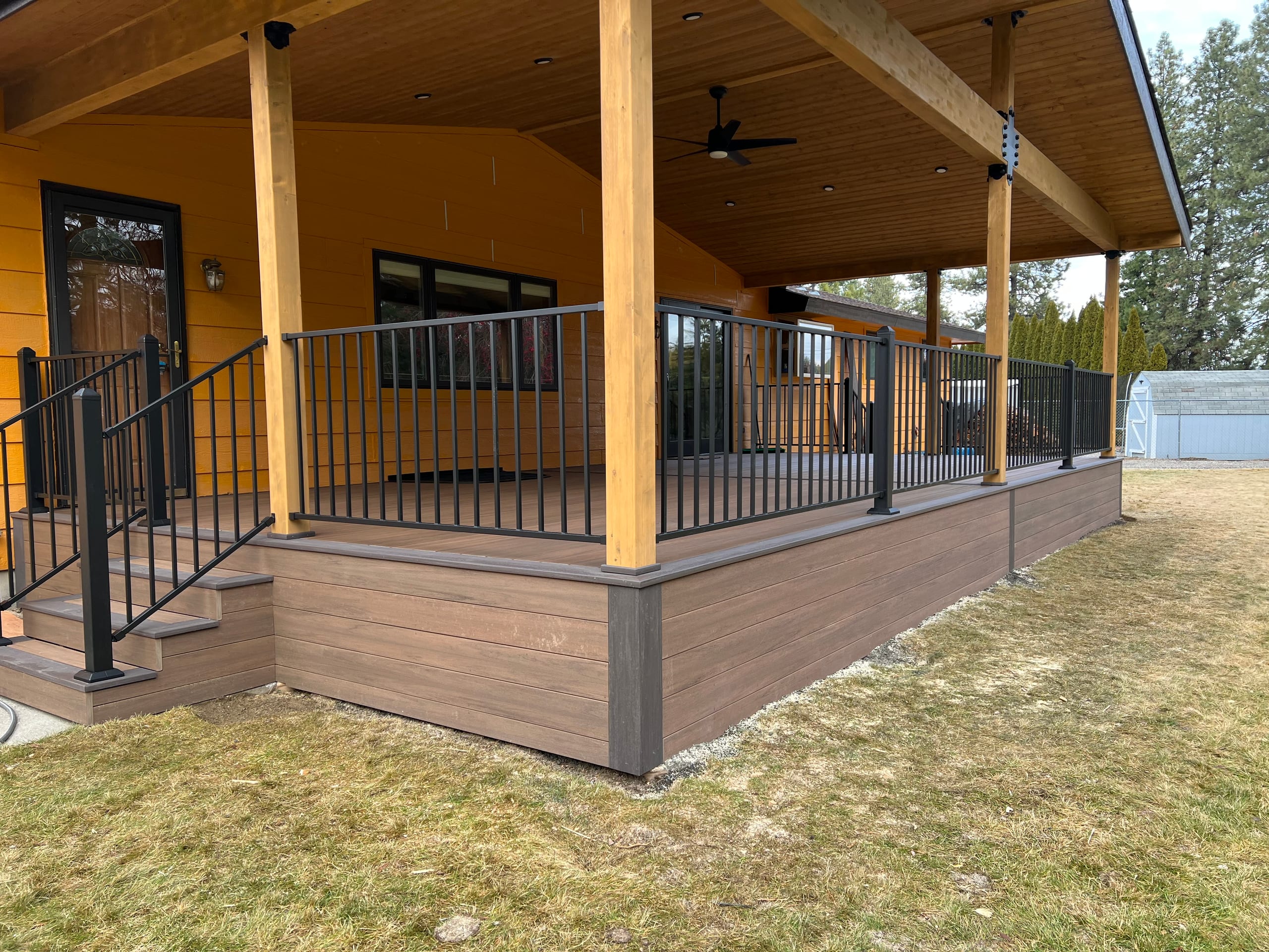 Corner of covered deck with cedar posts, black picket railing, steps and brown composite with grey trim on orange house
