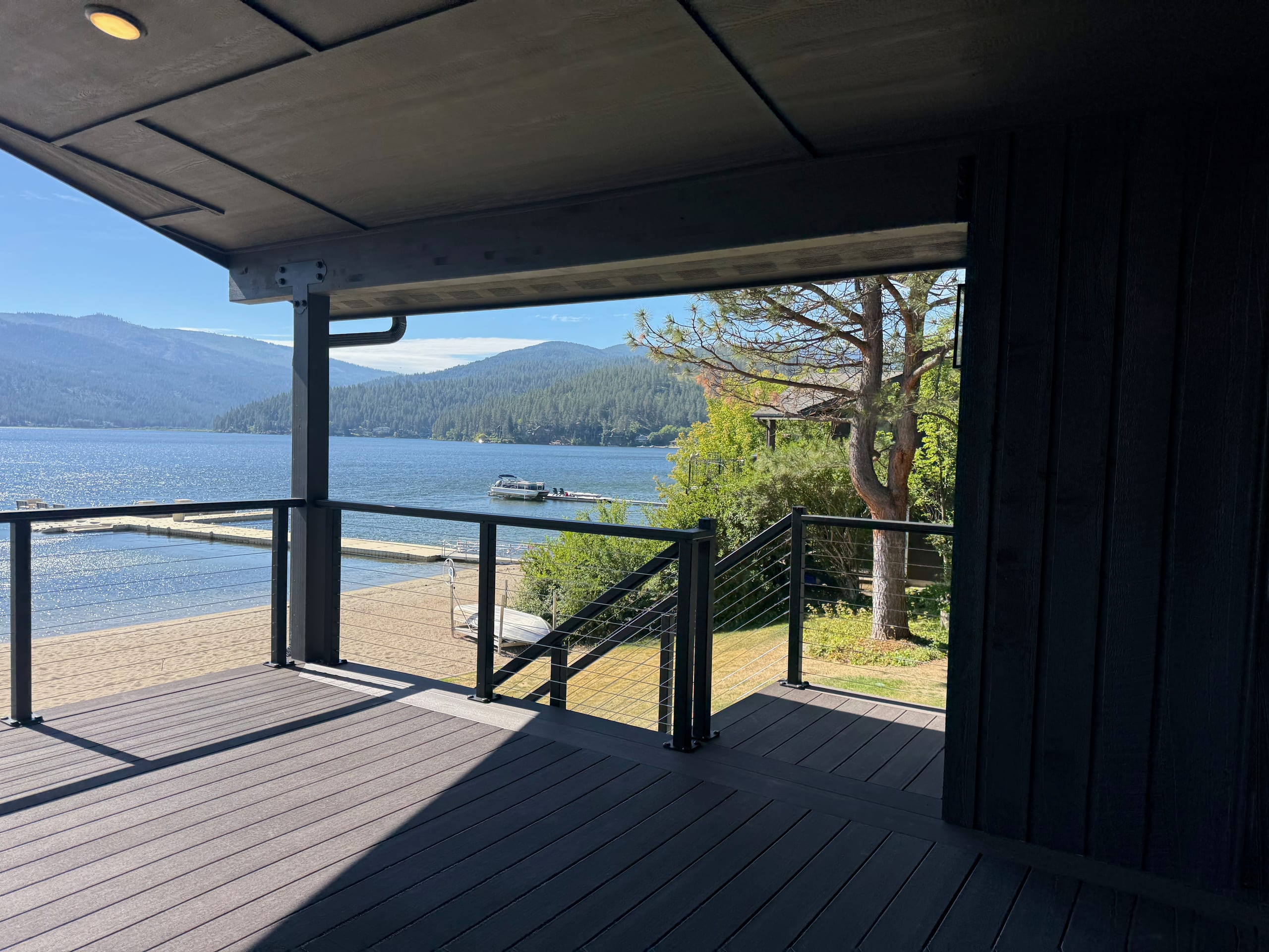 Corner of covered lakefront deck with cable railing and stair, dark composite boards, lake and mountains beyond