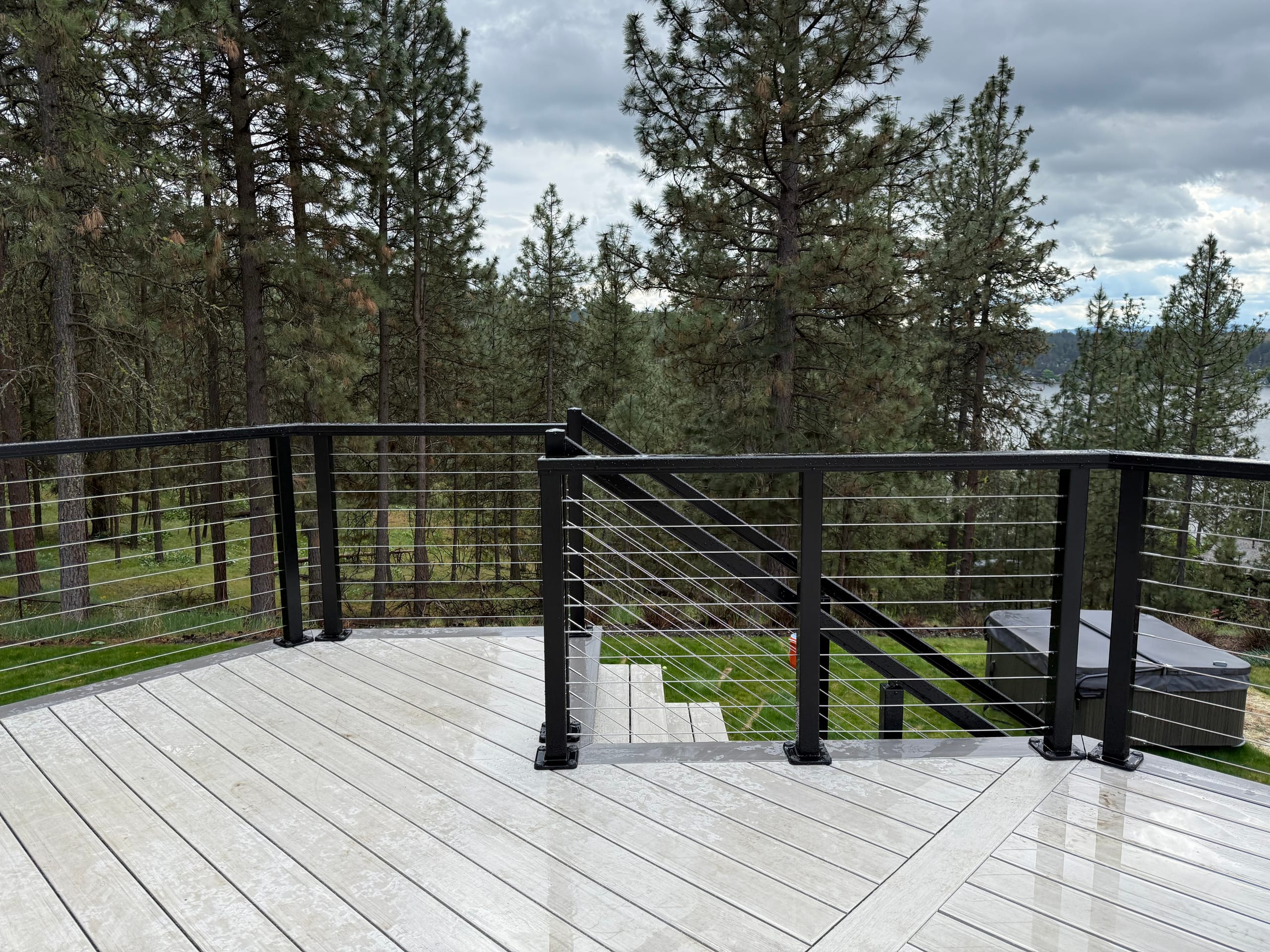 Corner of light gray composite deck with black cable railing and stairs, pines and lake glimpse
