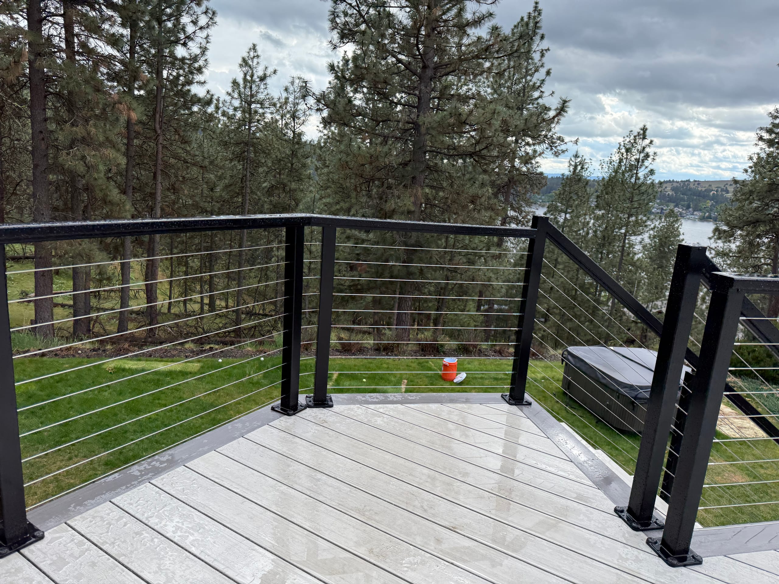 Corner of light gray composite deck with black cable railing overlooking lawn, hot tub, pines and distant lake