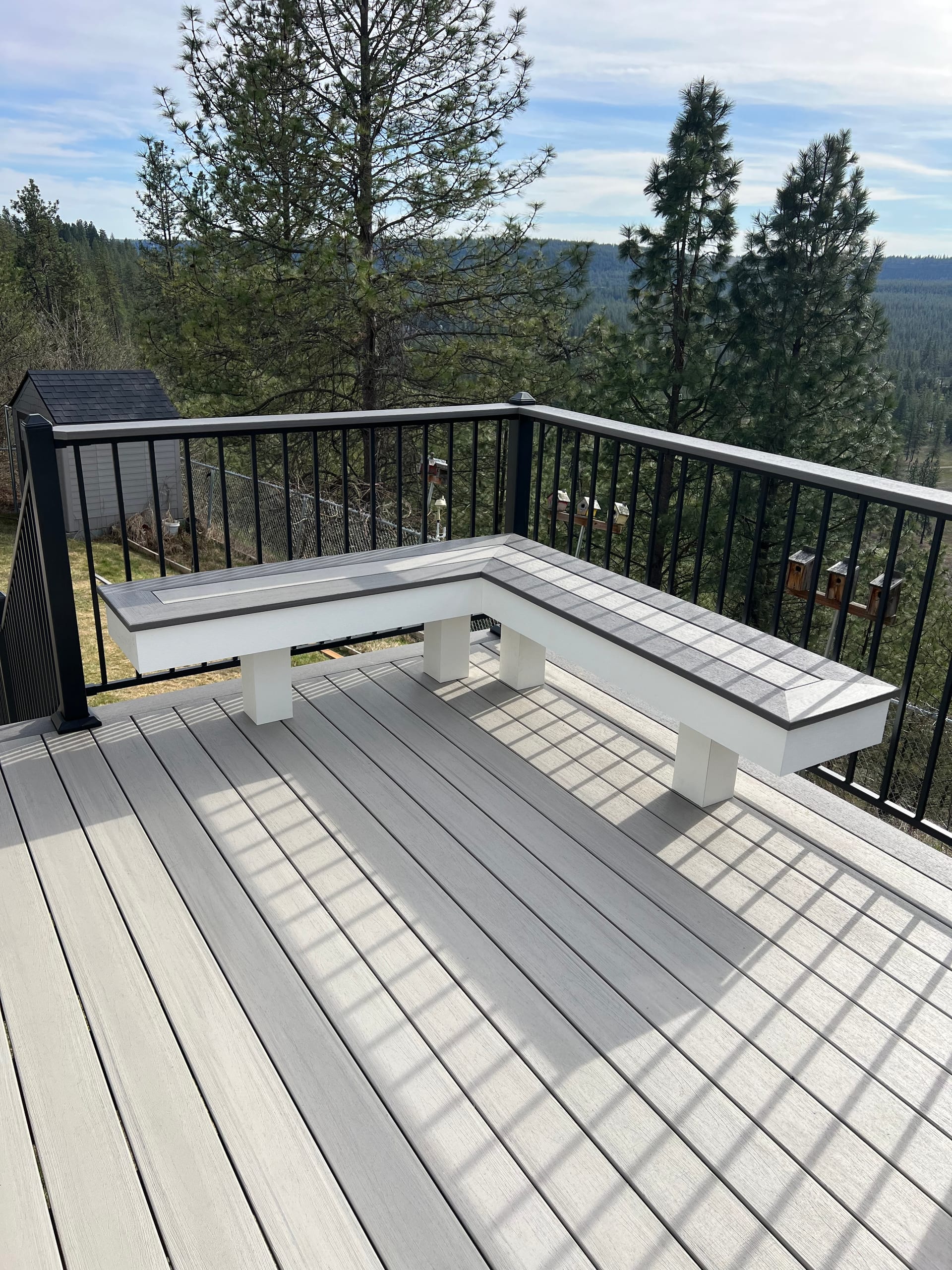 Corner of light grey composite deck with white L-shaped bench, black railing and hillside pine view