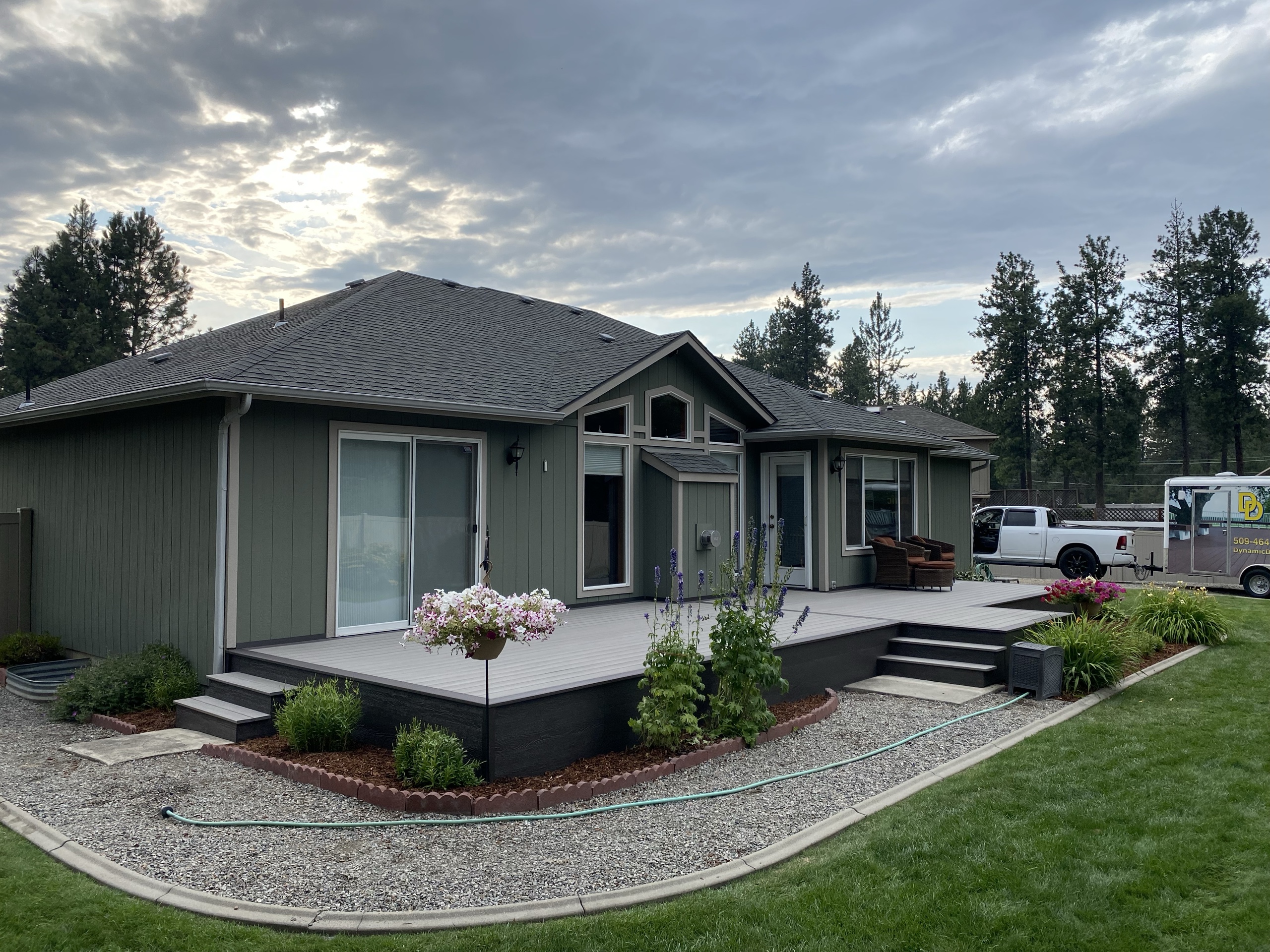 Corner view of low grey composite deck with steps, gravel edging and flowers on a green home, evening clouds