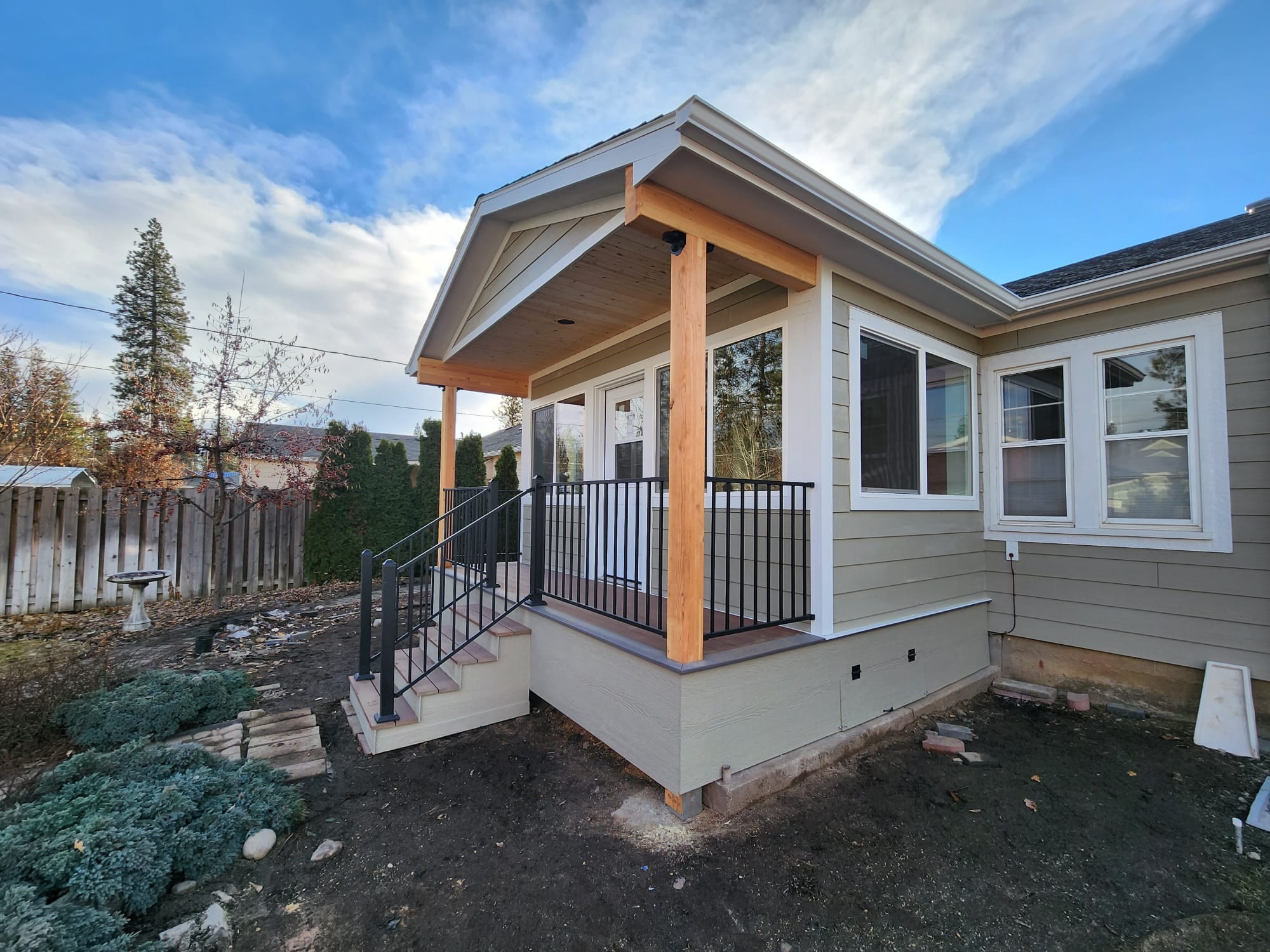 Corner view of a new enclosed room with gabled entry porch, stained timber posts, black railing and steps on a gray house