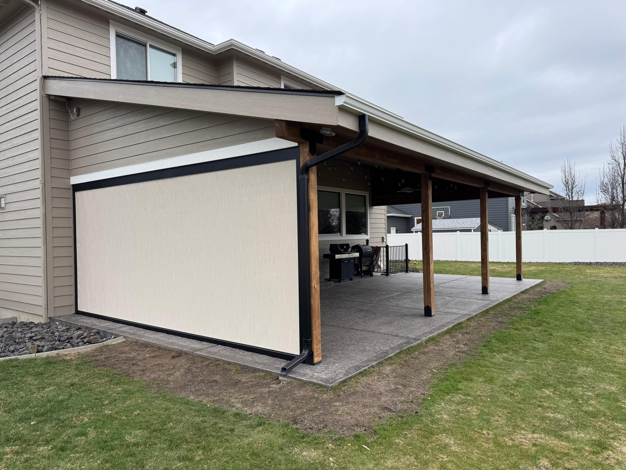 Corner view of patio cover with a roll-down privacy shade on one side over stamped patio, tan two-story house