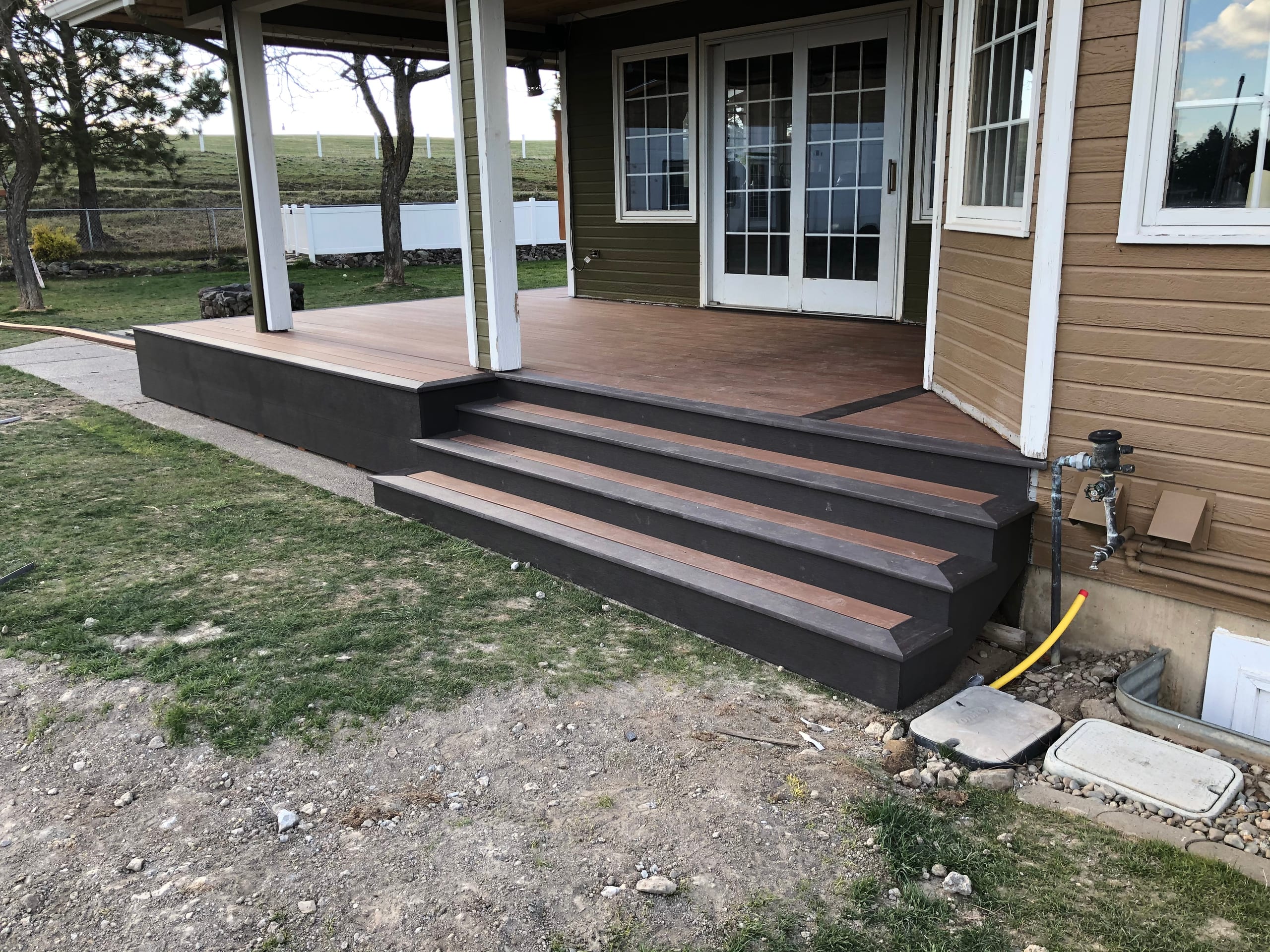 Covered back porch with dark brown composite deck and wide steps on a tan house with french doors, dirt yard