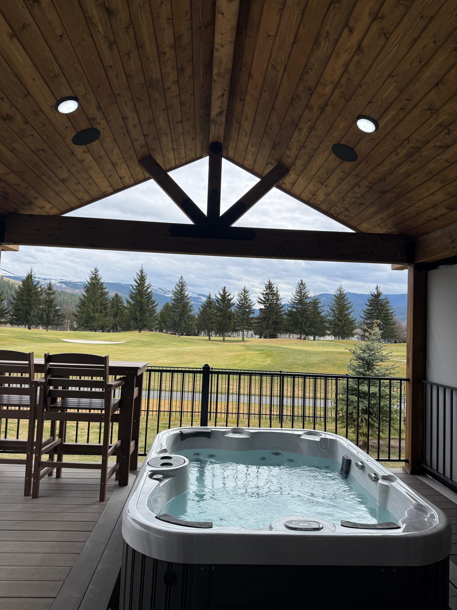 Covered deck with cedar gable ceiling, spa hot tub and dining table, black metal railing, golf course and mountains beyond
